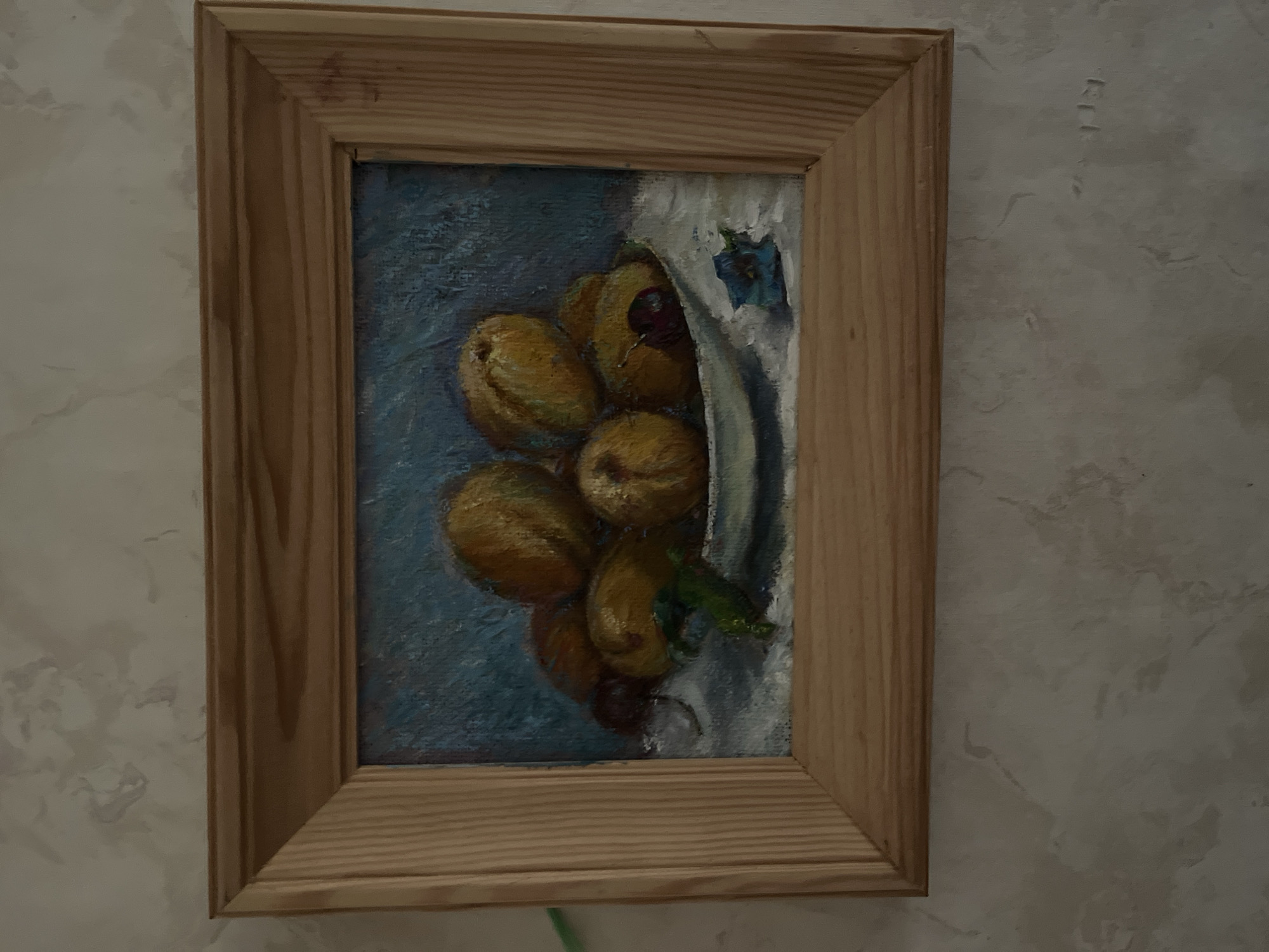 The media file is a vertical, medium shot photograph of a framed painting. The painting itself is a still life of several peaches in a white bowl, resting on a blue surface. A dark red fruit, possibly a cherry, is visible among the peaches, and a green leaf is also present. A portion of a white cloth is draped behind the bowl and peaches, and a small blue floral motif is painted on it. The painting is housed in a simple, light-colored wooden frame. The background of the photograph shows a textured, light-colored wall, possibly wallpapered or plastered, with a slightly mottled pattern. The lighting suggests an indoor setting, with the light source possibly coming from the left and slightly above. There are no people visible in the image, and no specific time of day or weather conditions can be determined. The location context of Starye Politsy, Russia, is provided externally and is not discernible from the image itself. No text is visible within the painting or the frame.