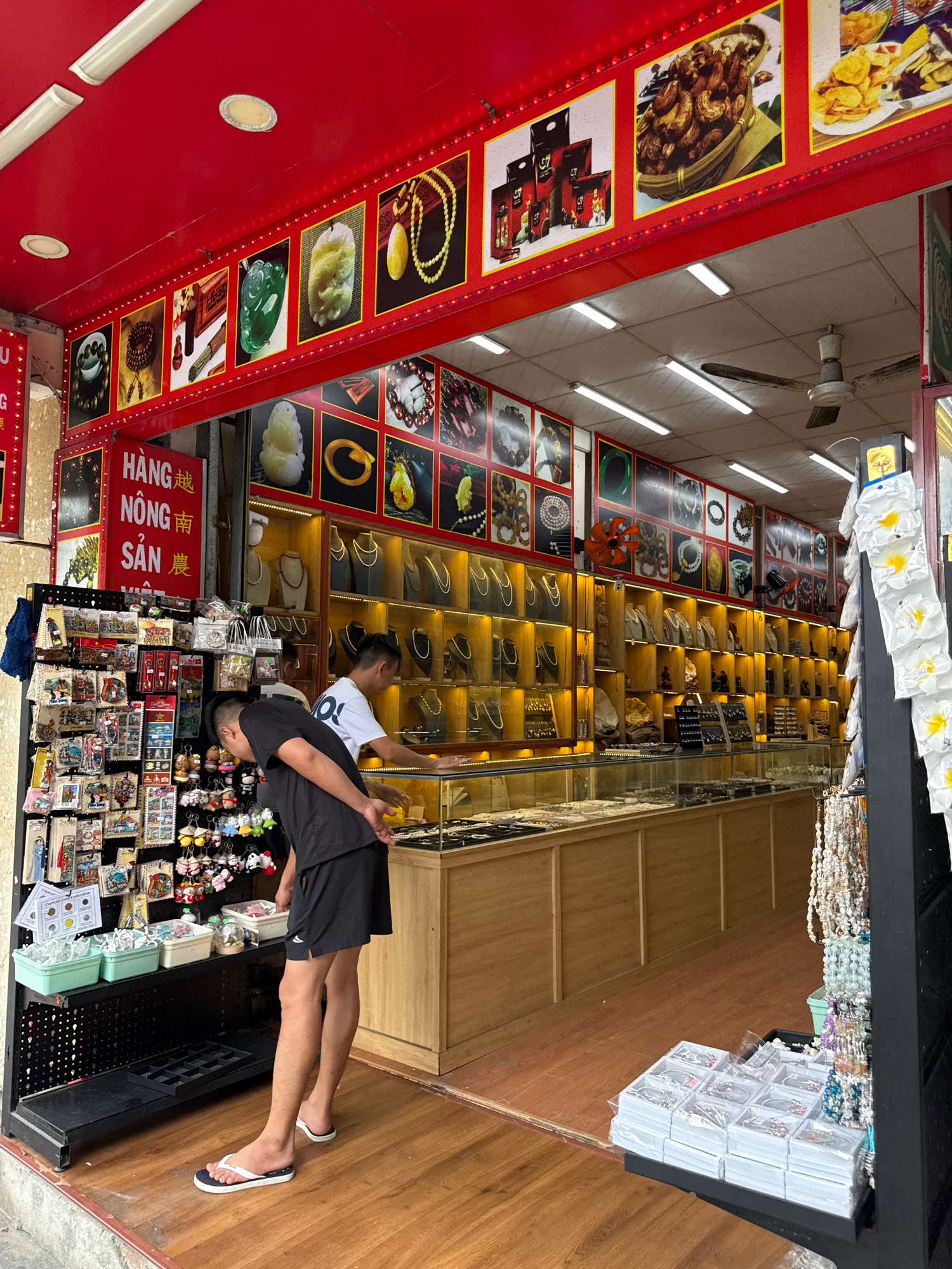 This image shows the interior of a shop in Nha Trang, Vietnam, specializing in jewelry and local handicrafts. The shop is brightly lit with fluorescent lights and decorated with a vibrant red awning and signage. The walls are adorned with framed pictures of jewelry, food items, and traditional Vietnamese art. In the foreground, two young men are browsing the merchandise. One man, wearing a black t-shirt and shorts, is looking down at a display of small trinkets. The other man, in a white t-shirt, is standing behind a glass display case filled with necklaces. The display cases themselves are made of wood and glass, and are filled with an array of necklaces, bracelets, and other jewelry items. In the background, shelves are stocked with various goods, including what appear to be souvenirs and local food products. The floor is made of polished wood. The overall impression is of a busy, well-stocked shop catering to tourists.