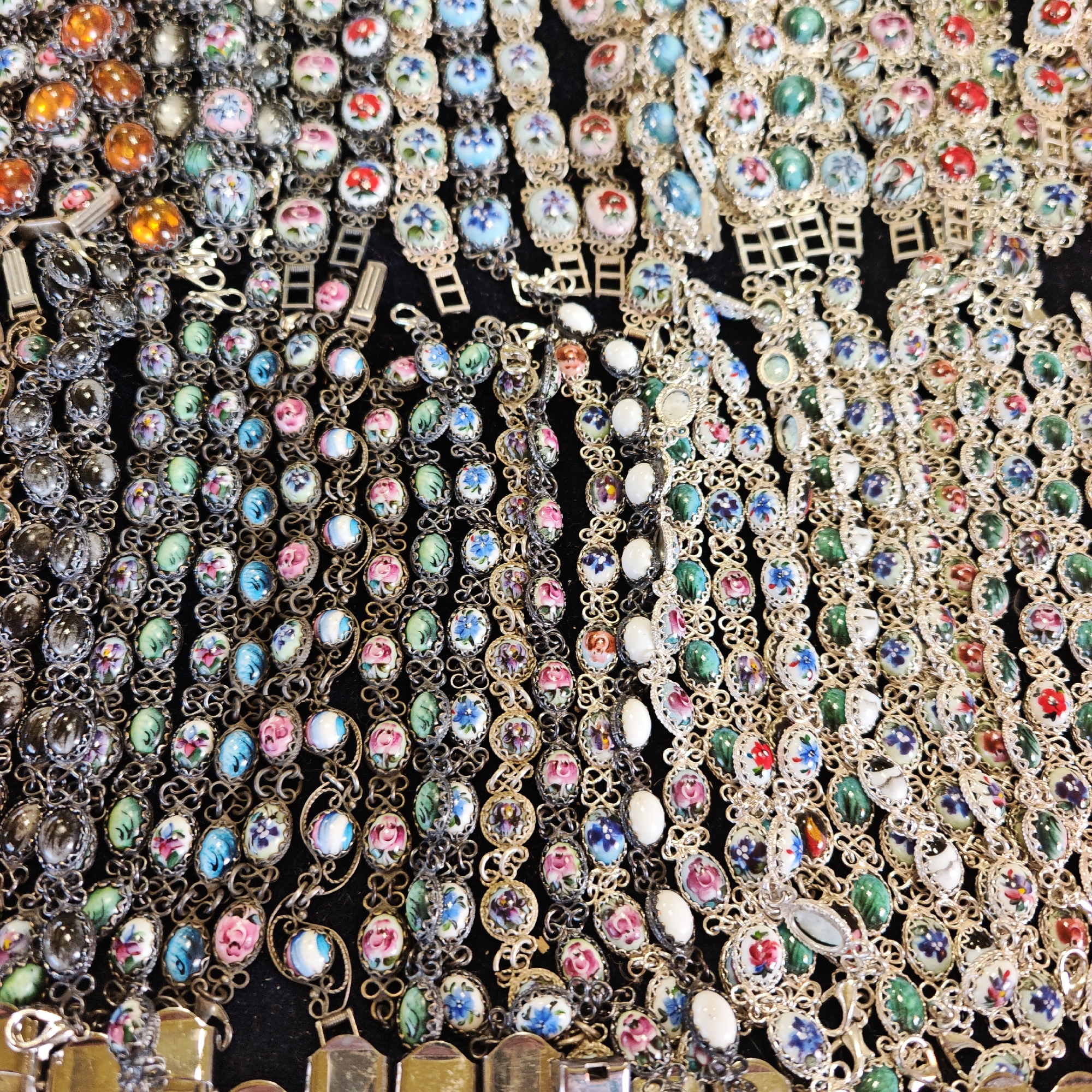 The image displays a close-up view of numerous bracelets arranged on a dark surface, likely a display at a market or shop. The location appears to be Unknown, Unknown. The bracelets are crafted with oval or circular links, each containing a decorative element - some are painted with floral designs in various colors, while others feature solid-colored stones or cabochons in hues like orange, green, and white. The metal settings appear to be silver-toned. The bracelets are entangled and overlapping, filling the frame with their intricate designs. The focus is on the details of the jewelry, with the dark background serving to highlight the colors and textures of the bracelets.