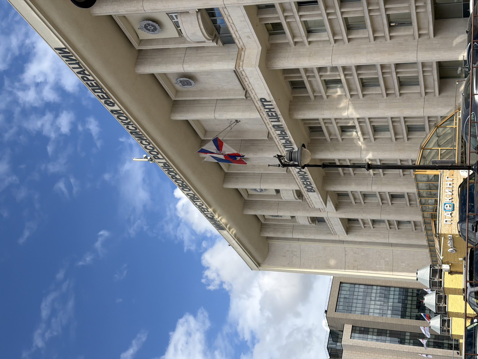The image shows the Ministry of Defence of the Russian Federation building in Moscow. The building is a large, rectangular structure made of light-colored stone. It has a prominent overhang with lettering that reads "МИНИСТЕРСТВО ОБОРОНЫ РОССИЙСКОЙ ФЕДЕРАЦИИ" (Ministry of Defense of the Russian Federation) and "ВОЕННЫЙ ЦЕНТР" (Military Center). A Russian flag is visible hanging from the building. A lamppost is in the foreground. A parking garage is visible to the right, and a modern building is situated in the lower portion of the image. The sky is blue with scattered clouds.