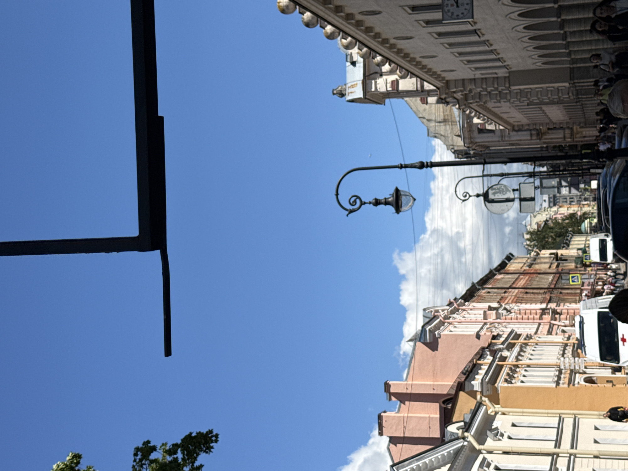 The image shows a street scene in Saint Petersburg, Russia, with a view looking up at buildings and the sky. The buildings feature ornate architecture, typical of the city, with detailed facades, arched windows, and decorative elements. In the foreground, a dark, angular structure, possibly a balcony or overhang, partially obscures the sky.
Along the street, there are lampposts with decorative lights, and what appears to be a clock face on one of the buildings. People are visible on the sidewalks, suggesting a bustling urban environment. A white vehicle with a red cross emblem, resembling an ambulance or emergency vehicle, is parked on the street, and a traffic sign is also visible.
The sky is a clear, bright blue with some fluffy white clouds, indicating fair weather. The time of day appears to be daytime, likely late morning or early afternoon, given the bright sunlight. The overall impression is one of a lively and historic city on a pleasant day. There is no visible text in the image, other than what might be on the traffic sign which is not legible.