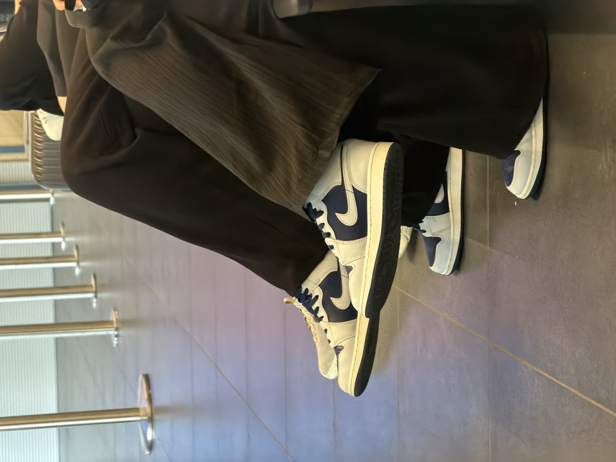 The image shows a person seated with legs crossed, wearing black trousers and what appears to be a dark grey or black textured top. The person is wearing white and blue Nike Air Jordan 1 high-top sneakers. The background shows a floor with a dark gray surface and a queuing system with metal poles. The setting looks like an airport or a large public space. There is no definitive evidence to suggest the image was taken in Budapest, Hungary.