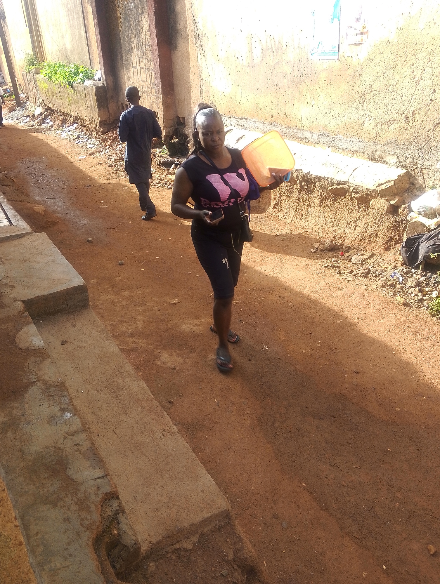 The image depicts a street scene in Jos, Nigeria. A woman in a black t-shirt and shorts is walking down a dirt road carrying an orange bucket. A man in a dark blue outfit is walking in the same direction a short distance behind her. The road is lined with concrete structures and walls, some covered in graffiti or vegetation. Debris is scattered along the roadside. The lighting suggests it is daytime, with strong shadows present.