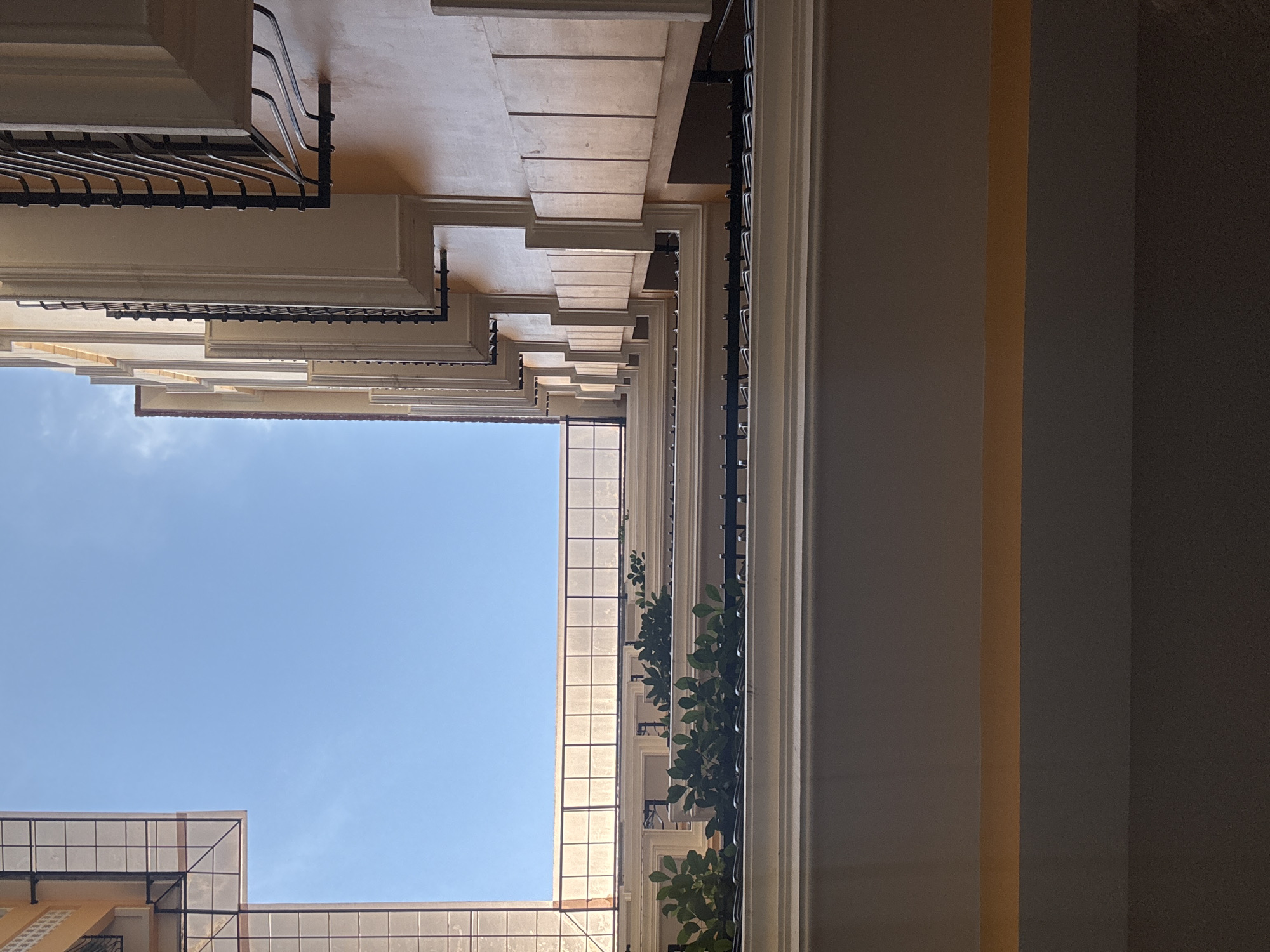 The image presents an upward-looking view from within a narrow urban architectural space, possibly an interior courtyard or passage between buildings, towards a clear blue sky.
On the left side, a multi-story building facade is visible, characterized by a series of projecting balconies or ledges on each floor. These light-colored structures are equipped with dark metal railings featuring both decorative wavy elements at the top and a grid pattern below. The building's walls are a warm, light cream or beige, illuminated by bright daylight.
To the right, another section of the building rises, partially in shadow. This facade displays prominent light-colored vertical architectural features, possibly columns or pilasters. Dark metal grates or railings are affixed here, adorned with clusters of vibrant green foliage, suggesting climbing plants or vines.
Near the top, translucent panels with a grid pattern form part of a roof structure or skylight, allowing diffused light to penetrate the space. The central and upper portions of the image are dominated by a vast expanse of clear, pale blue sky, indicating bright, sunny weather, likely midday or early afternoon.
The very bottom right corner of the frame reveals a deeply shadowed area, appearing as an interior wall or a recessed part of the structure, featuring various shades of dark grey and a distinct vertical amber-brown stripe. The scene is devoid of people or specific activity, emphasizing architectural lines, light, and shadow. The architectural style and integrated greenery align with an urban setting like Đà Nẵng, Vietnam. No visible text is present.