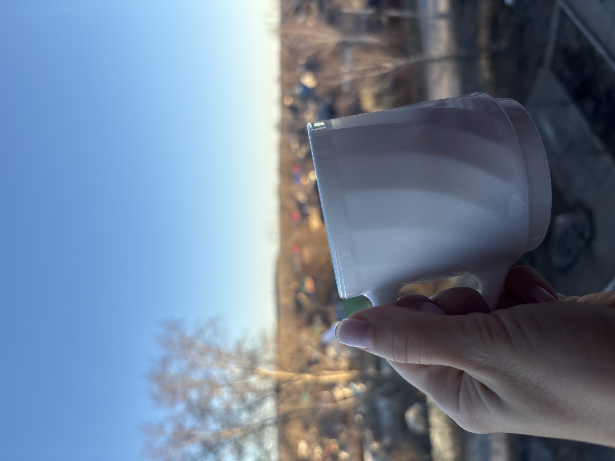 A hand holds a white mug against the background of a blurred cityscape. The sky is clear blue, visible to the left of the mug, and leafless trees are in the middle distance. The city buildings in the background are indistinct. The image was most likely taken from inside a building, possibly a home or office, based on the presence of a reflective surface in the lower right corner. The location appears to be the city of Chaikovsky, Russia, although specific landmarks are not clearly identifiable in this image.
