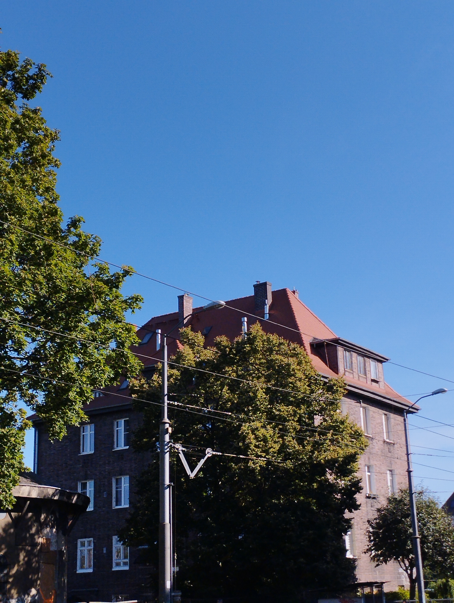 An overcast sky is visible in the background of the image, with a building and trees in the foreground. The building is made of brick and has a red tile roof. It has several windows, some of which are open. The trees are green and leafy. The image appears to be taken from a low angle, looking up at the building and trees. The image quality is poor, with the subject of the image being blurry and out of focus. This image was taken on the afternoon of June 19, 2023, at an unknown location in Szczecin, Poland.