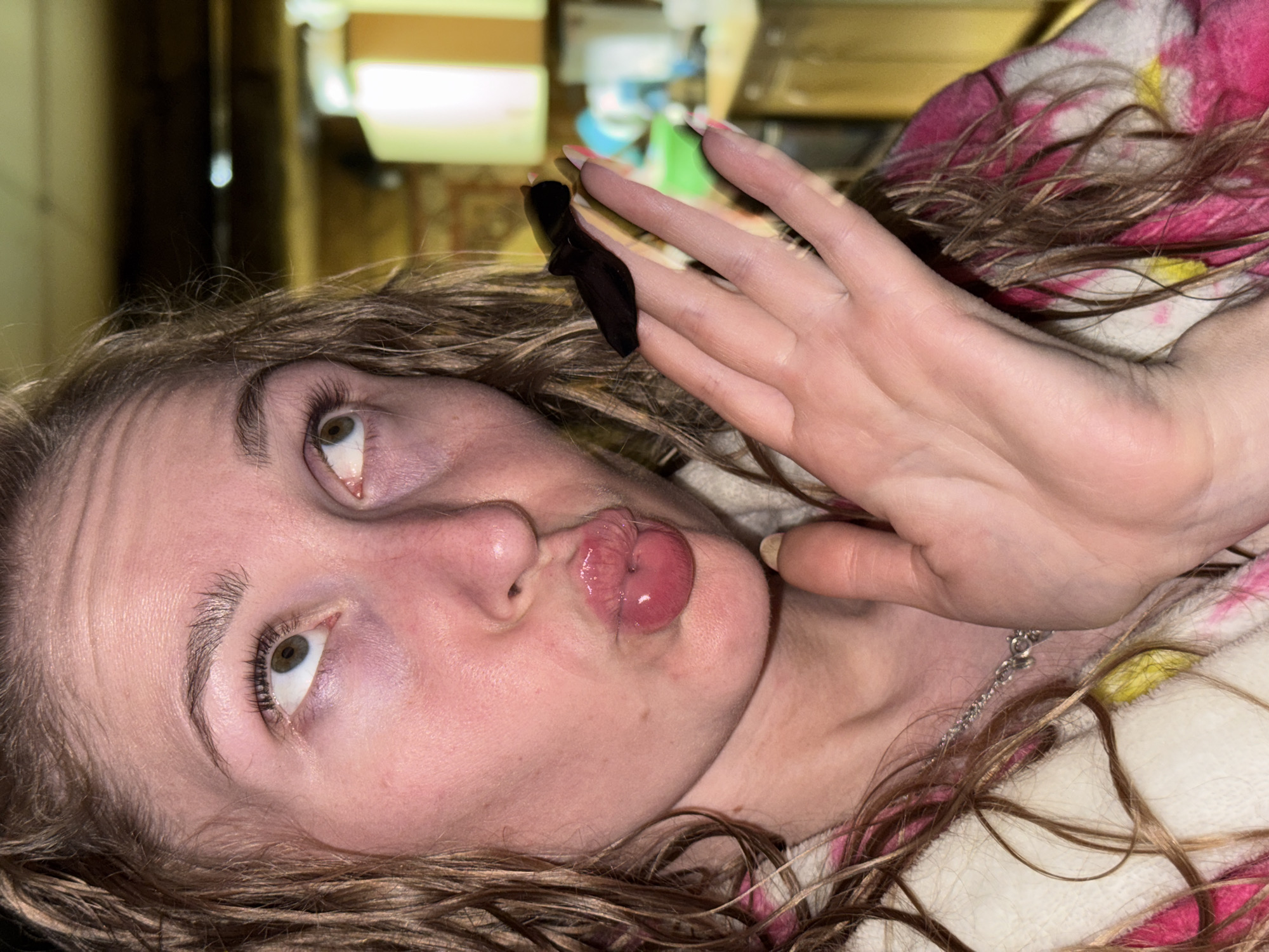 A person lies on their back with a pink and white patterned blanket partially visible. The person has light skin, brown hair, and is making a kissing face. One hand is raised with the palm facing the viewer. A black hair clip is visible in the hair near the hand. A silver necklace is around the person's neck. The background is blurry but appears to be an indoor setting. There is no evidence in the image to indicate it was taken in Nizhnyaya Chermoda, Russia.