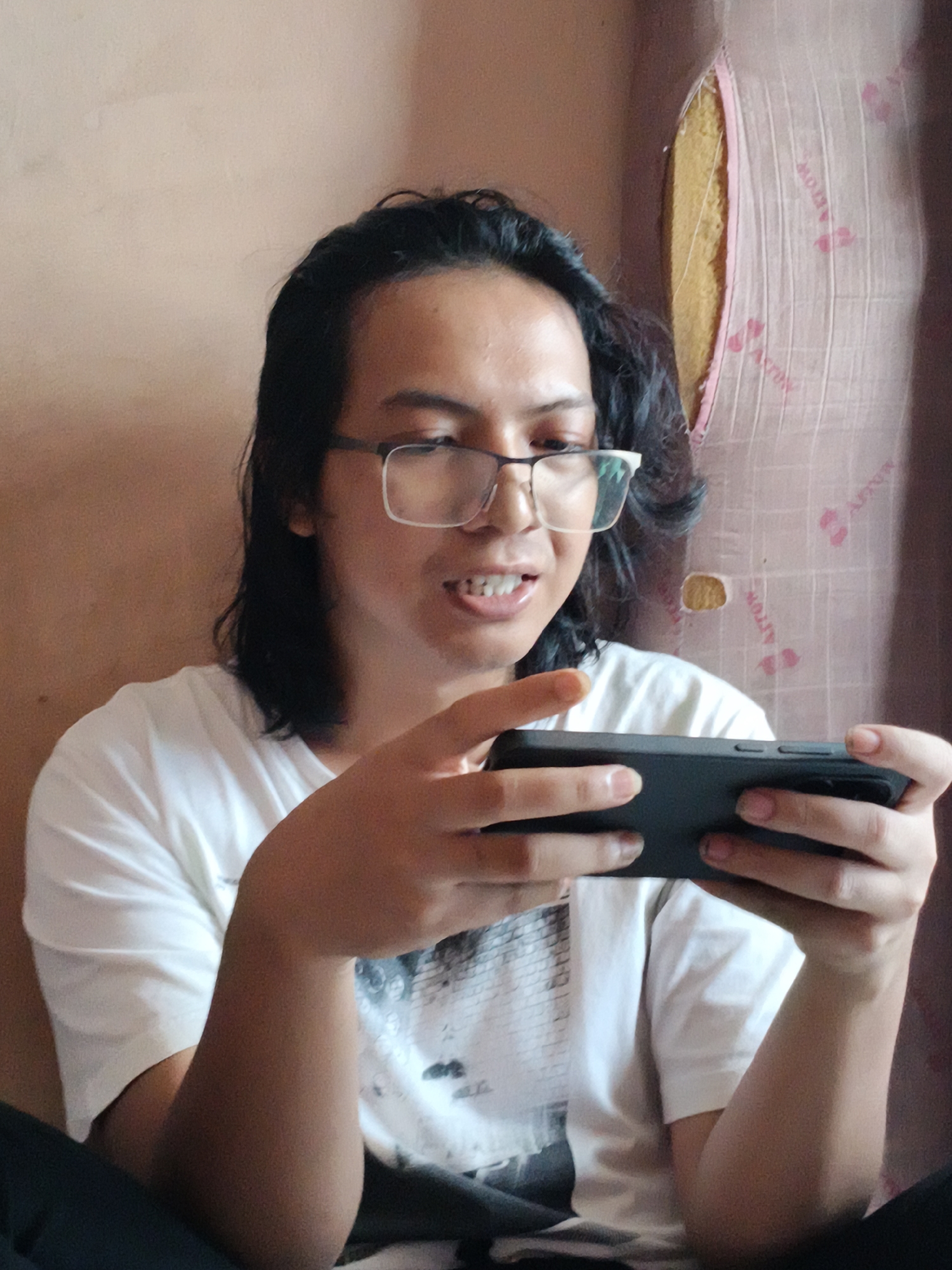 A person with long dark hair and glasses is sitting and holding a black smartphone in their hands, appearing to be interacting with it. They are wearing a white t-shirt. The background consists of a plain, light-colored wall and a torn, patterned cushion or padding. The location is likely indoors. The location is Cianjur, Indonesia.