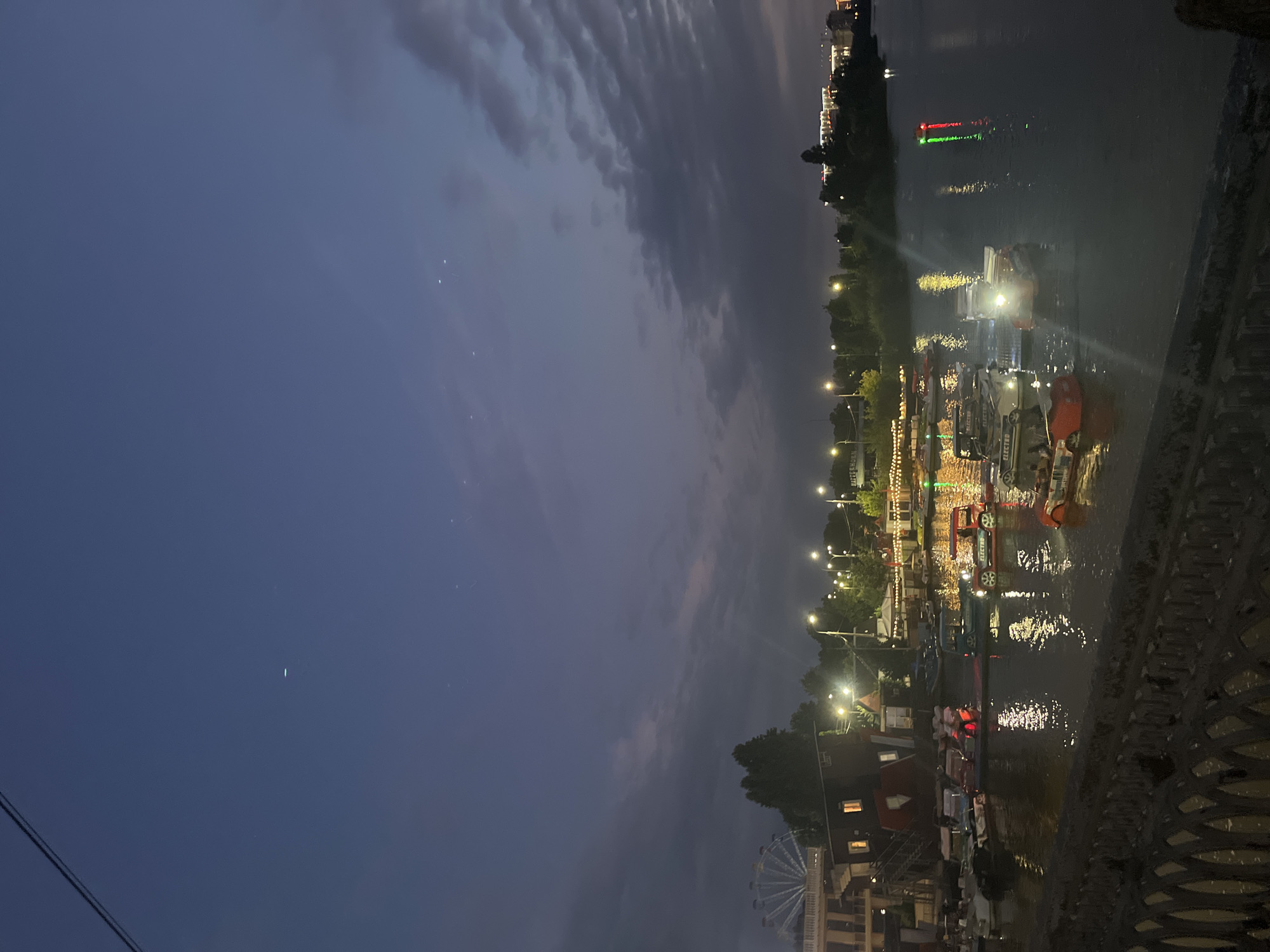 The image depicts a twilight scene at an amusement park or waterfront area in Krasnodar, Russia. The sky is a deep twilight blue with scattered clouds and a few visible stars. In the distance, a large Ferris wheel illuminated with white lights is visible on the left side of the frame, suggesting the presence of an amusement park.
The foreground features a body of water, likely a river or canal, with several small boats and pedal boats docked or moving on the surface. Some of these boats have lights on, reflecting on the water. One red pedal boat has the word "ELECTRO" printed on its side. The water is calm, with reflections of the lights from the shore and the boats.
Along the shore, there are buildings, trees, and streetlights. The buildings appear to be part of the amusement area, with some illuminated windows. The trees are silhouetted against the darkening sky. The overall impression is of a lively evening atmosphere with recreational activities taking place near the water.
The location is identified as Krasnodar, Russia, which suggests this scene is from an area along the Kuban River or a similar waterfront recreational zone within the city.