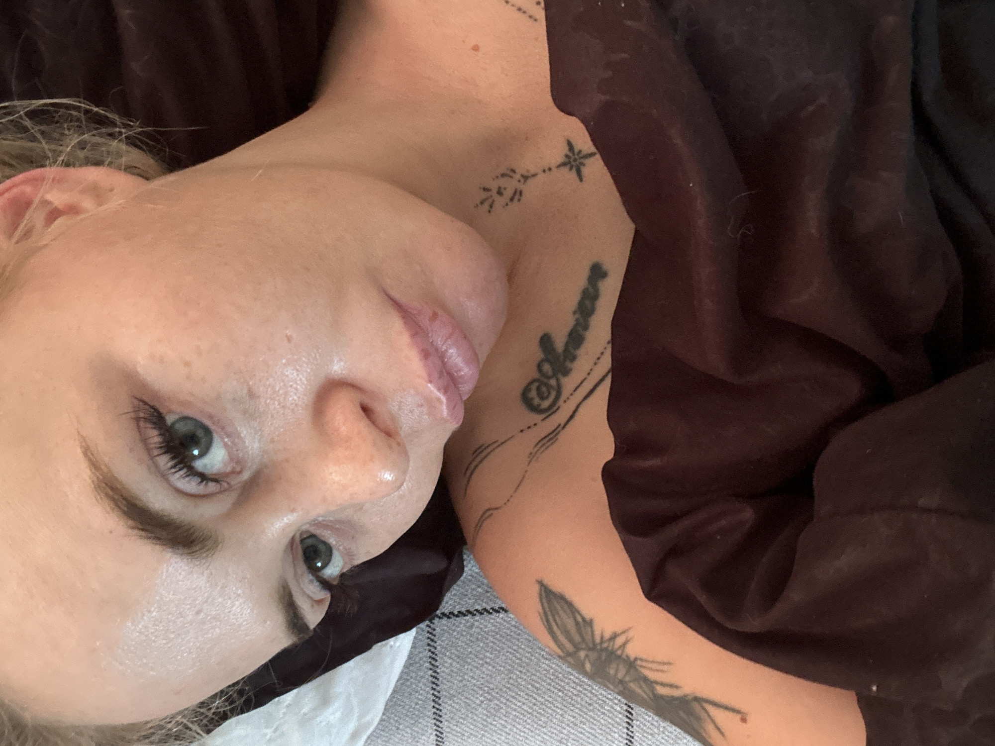 A young woman with blonde hair, green eyes, and light skin with freckles is lying down, looking at the camera. She has makeup on, including mascara and lip gloss. She has tattoos on her shoulder and chest. The woman is covered by a dark red blanket. The background is a grey and white checkered fabric. The setting is indoors, possibly a bedroom.