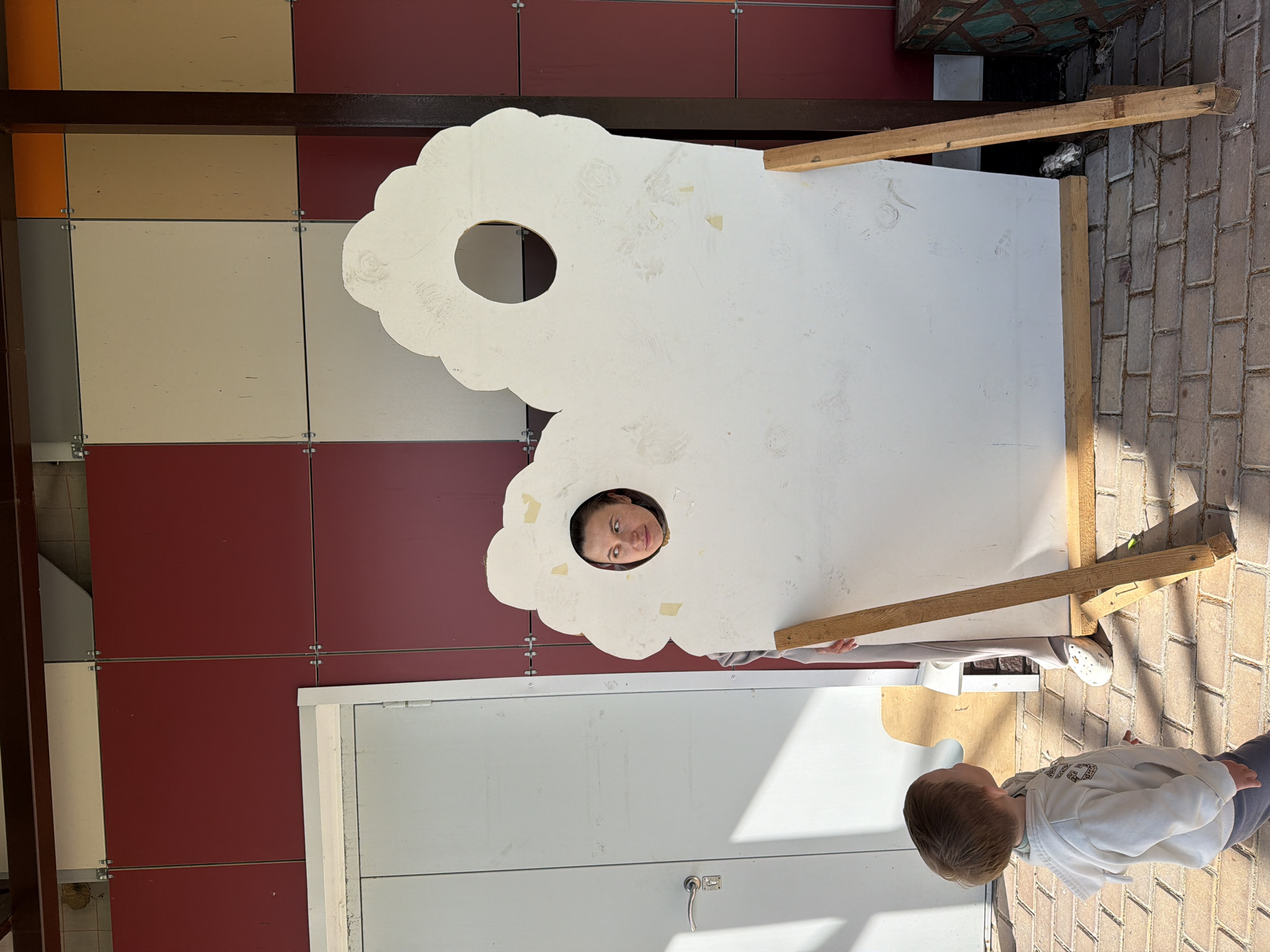 The image shows a young boy and a woman. The woman is peeking through a cloud-shaped cutout in a large white board. The boy is looking up at the woman. They are outdoors, with a building and brick pathway visible. The building has a colorful facade, with panels in shades of orange, beige, red, and grey. A white door is visible on the lower part of the building. The lighting suggests it is daytime, with shadows cast on the ground. The scene appears to be a playful or educational setting. There is no visible text in the image.