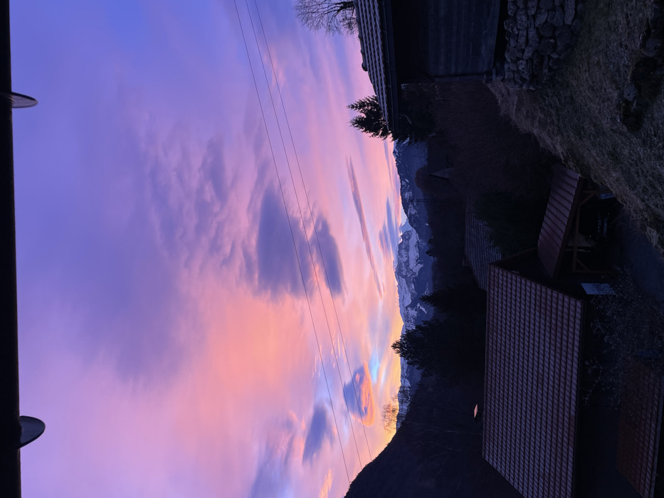 The image captures a serene mountainous landscape at either sunrise or sunset, dominated by a spectacularly colored sky. The upper portion of the frame is filled with vibrant hues of purple, pink, orange, and pale yellow, interspersed with clouds ranging from dark and defined to wispy. Several power lines or cables stretch horizontally across the sky.
Below the dramatic sky, a range of mountains unfolds, featuring distinct snow-capped peaks in the distance and darker, tree-covered foothills closer to the viewer. Several buildings, possibly houses or barns, with dark or reddish-brown tiled roofs, are visible in the midground and foreground, nestled within the landscape. A lamppost is partially visible on the far left, and patches of dry grass and a stone wall are also discernable.
The time of day is clearly marked by the intense, glowing colors of the sky, indicating either dawn or dusk. The low light casts parts of the foreground and midground elements, such as the buildings and closer trees, into partial silhouette. The overall mood is peaceful and majestic, showcasing the natural beauty of a mountainous region during a transitional light period. There is no specific activity taking place. No visible text is present in the image.