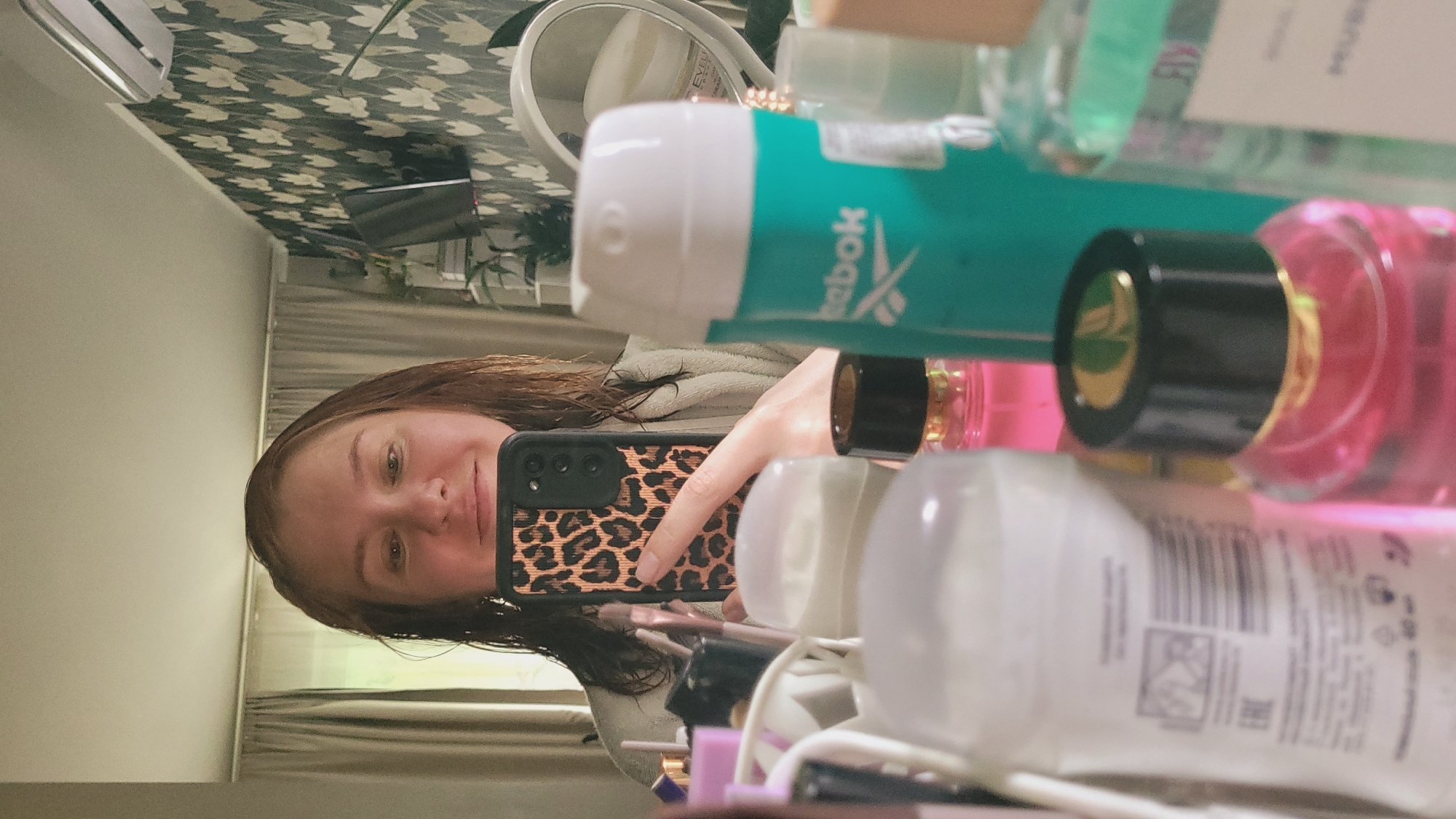 The image is a selfie taken in a mirror, showing a woman with brown hair and a light-colored robe. She is holding a smartphone with a leopard-print case to take the picture. Various cosmetic products are visible on a shelf or surface in front of her, including bottles with colorful liquids and opaque white containers. Some of these bottles have labels, though the text is largely illegible. One teal-colored bottle has the Reebok logo visible. The background includes a patterned wallpaper, a mirror, and curtains. The lighting suggests it might be daytime. The woman appears to be smiling, and the overall mood seems casual and relaxed.