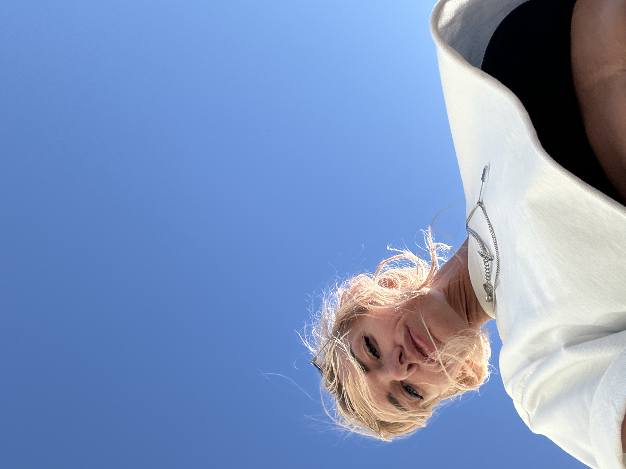 A low-angle, close-up shot features a woman with short, blonde hair and pink undertones against a bright, clear blue sky. She is looking down towards the camera, her face partially obscured by her hair, which is windswept. She wears a white t-shirt with a silver chain necklace. The background is entirely composed of the sky, suggesting an outdoor setting. There are no discernible elements that would indicate a specific location within Saint Petersburg, Russia.