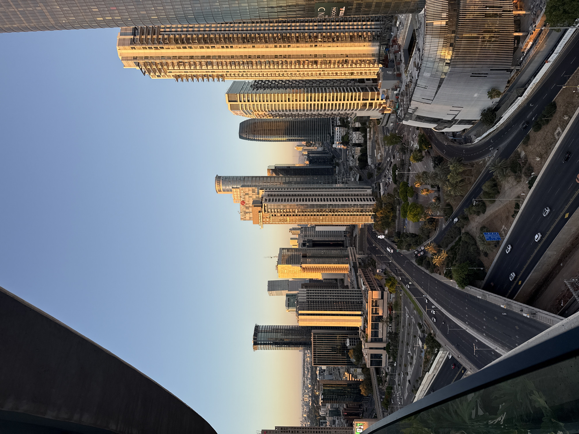 This is a high-angle, long shot photograph of the skyline of Tel Aviv, Israel, at sunset. The image is dominated by tall modern buildings, many of which are illuminated by the warm, golden light of the setting sun. The foreground features a curved, dark gray balcony railing and a sliver of green foliage, suggesting the viewpoint is from a high-rise building.
The cityscape extends into the distance, with a clear blue sky above. Several multi-lane highways and roads weave through the urban landscape, with a few cars visible on them, indicating moderate traffic. The roads appear to be elevated in some sections, crossing over one another. Palm trees and other greenery are interspersed between the buildings and along the roadsides, adding a touch of nature to the urban environment.
One prominent building in the mid-ground features a unique, curved facade with vertical ribbed elements, appearing to be under construction or recently completed. Other buildings are a mix of glass, concrete, and metal, showcasing contemporary architectural styles. The overall impression is one of a vibrant, modern city bathed in the beautiful light of a sunset. The specific location is identifiable as the Tel Aviv skyline, known for its modern skyscrapers and coastal setting.