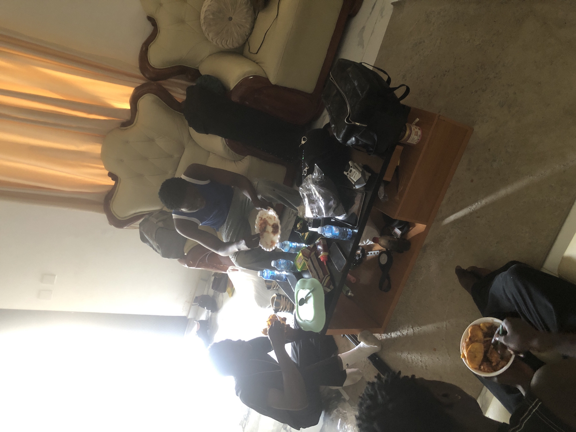 Three young Black men are gathered in a living room, likely in Ado, Nigeria, where bright sunlight streams in from a large window on the left. Two men are seated on the floor around a low wooden coffee table laden with food and drinks. The man on the left, wearing a black t-shirt and white socks, is eating from a plate of food. Another man, seated closer to the table, is also eating from a plate. The third man is seated on a plush, cream-colored sofa to the right, also appearing to be eating.
The coffee table holds several bottles of water, a bowl of food, snacks, and a tablet. Bags, including a black backpack, are scattered around the room. The room is furnished with ornate sofas and curtains. The floor is a tiled surface, and the walls are a light color. The overall impression is a casual gathering, possibly a mealtime, within a domestic setting.