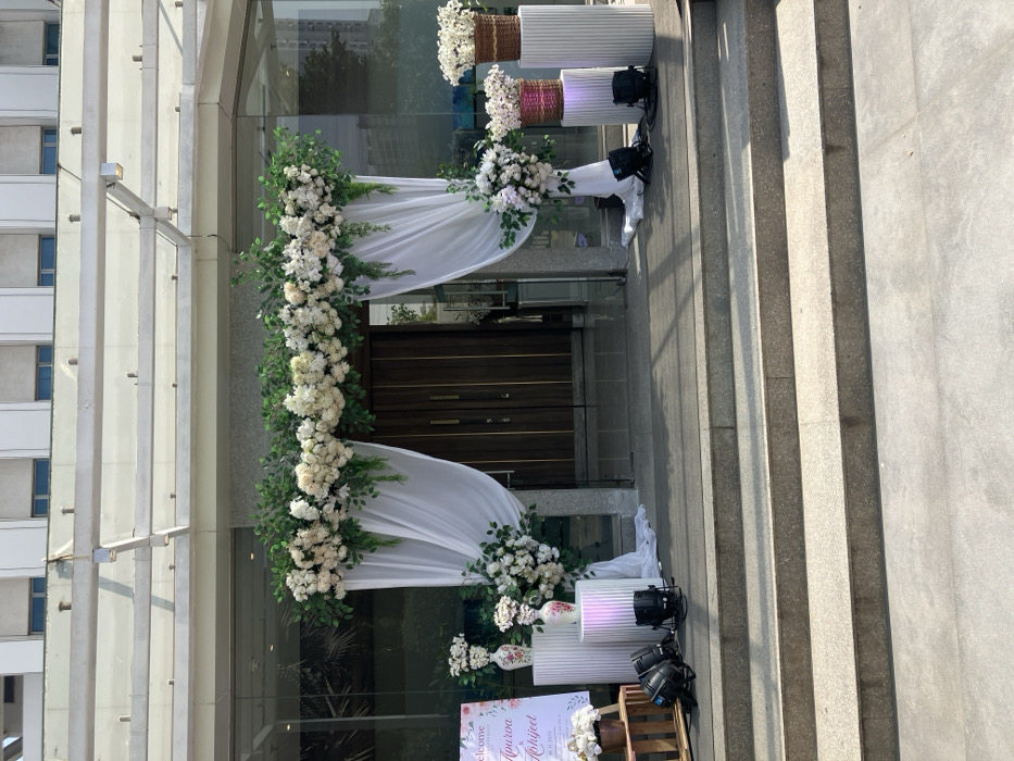 An exterior shot reveals a building adorned for an event. A floral archway, lush with white flowers and greenery, frames the entryway doors. White fabric is draped elegantly. Further decorations include cylindrical white stands, some topped with floral arrangements in baskets or vases. Lighting equipment is visible near the stands. Steps lead up to the decorated entrance, all set against the backdrop of the building's architecture.
