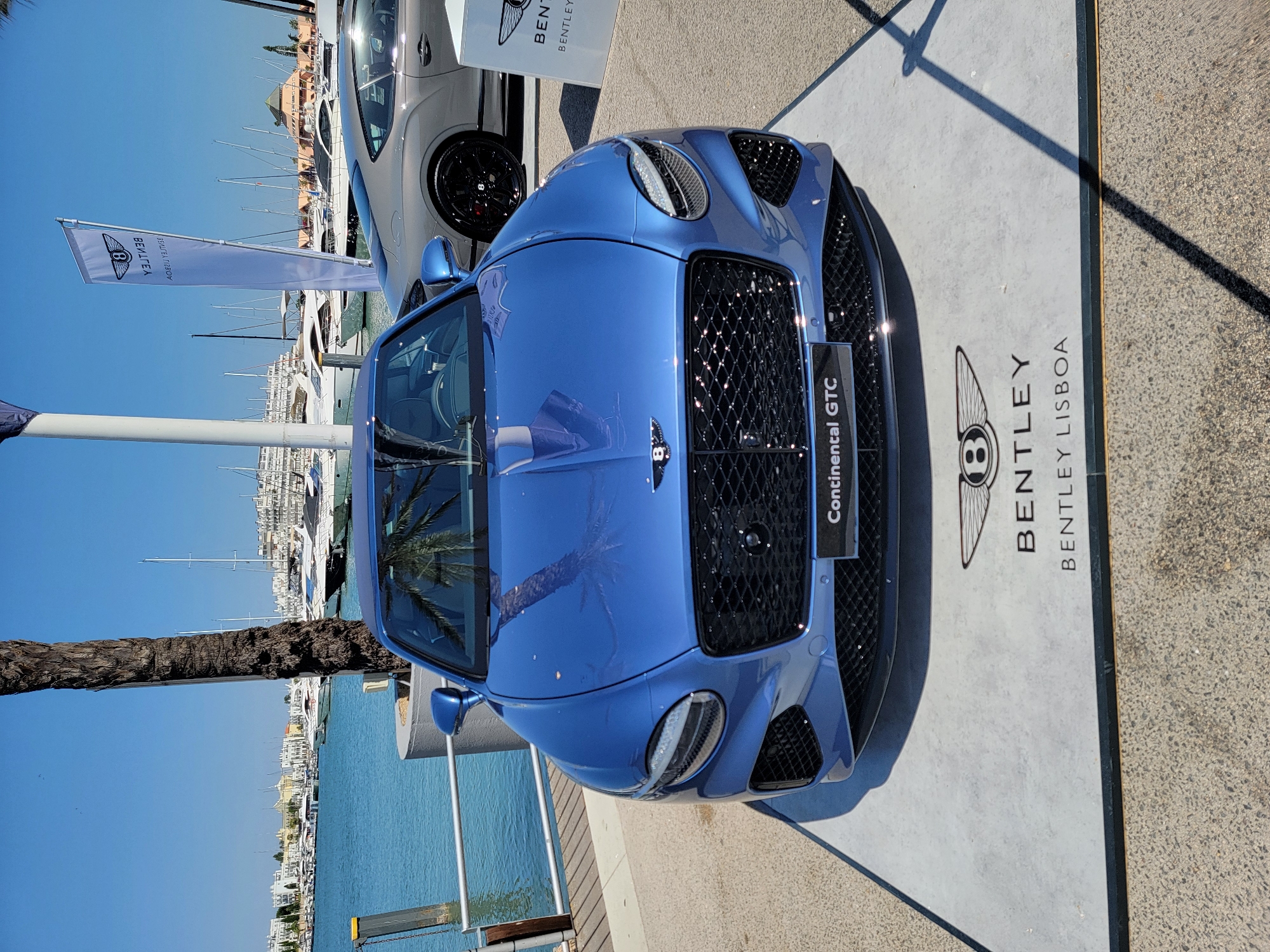 The image shows a blue Bentley Continental GTC parked on a display mat with the Bentley logo and "BENTLEY LISBOA" written on it. The car is positioned in the foreground, with its front grille and headlights clearly visible. Behind the blue Bentley, a silver Bentley is parked, and further in the background, there is a marina filled with yachts and boats. The scene is set in a marina in Quarteira, Portugal, evident from the distinct architecture of the buildings along the waterfront and the clear blue sky. A tall, thin flag with the Bentley logo is also visible to the right of the car. The overall atmosphere is that of a car exhibition or dealership event taking place in a scenic waterfront location. There are no people visible in the image.