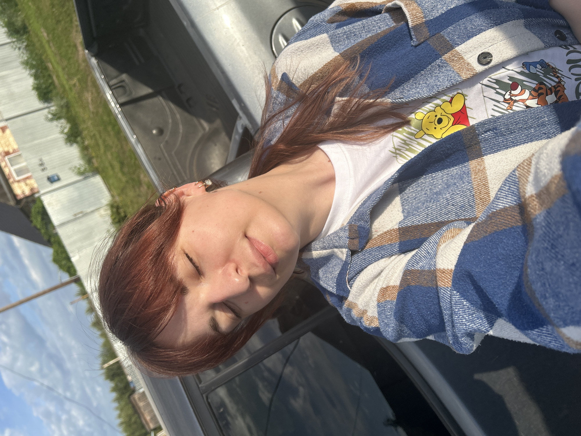 A woman with auburn hair is depicted close-up, eyes closed. She is wearing a white t-shirt with a Winnie the Pooh design and a blue and tan plaid flannel shirt. She appears to be leaning against a silver vehicle. Outside the vehicle window, a partly cloudy sky, a building, and greenery are visible. The location Фоки, Russia is not distinguishable from the image.