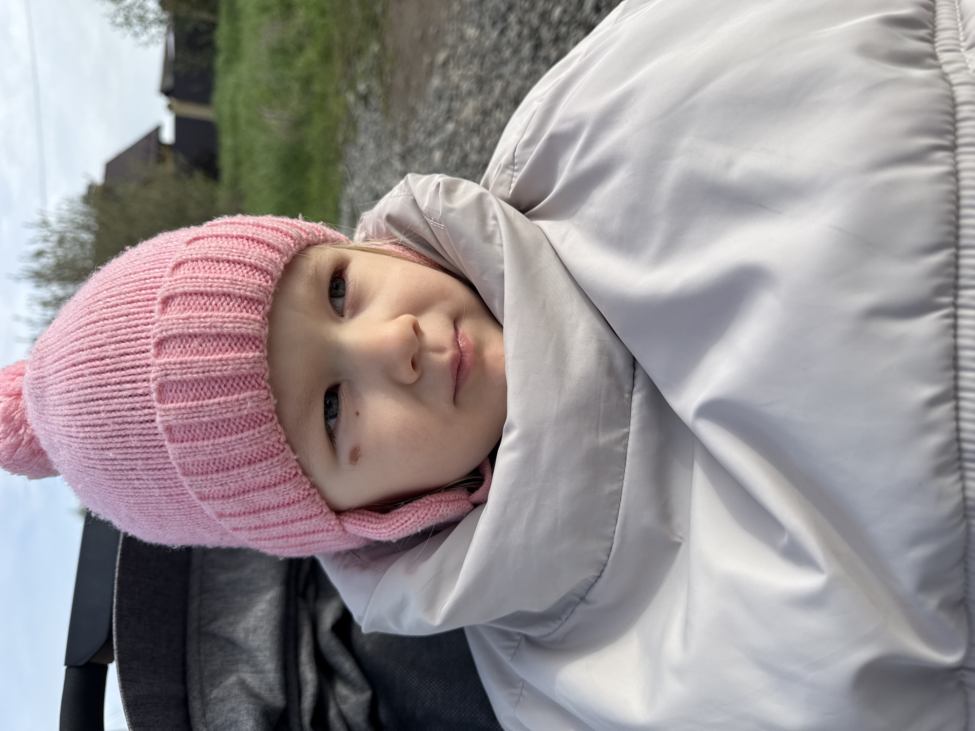 A close-up, medium shot depicts a young child wearing a pink knit hat and a light grey, puffy coat with a hood, positioned at an angle. The child's face is visible, displaying a calm expression. The background includes a gravel surface and glimpses of greenery and buildings. The location is Горный, Russia.