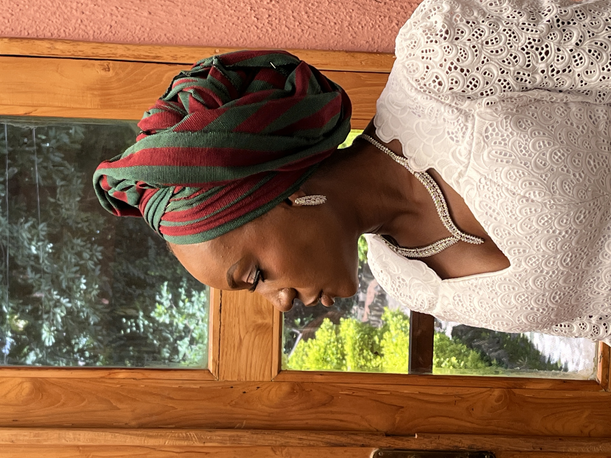 A woman, viewed from the side in a profile shot, gazes out a window. She is wearing a green and red striped headwrap, a white lace dress, and a diamond necklace and earrings. The woman is standing next to a wooden framed window, which looks out onto lush green foliage. The window is framed by a light brown wooden door. The wall to the right of the woman is a textured pink. The location is Bar Jirgi Summa, Nigeria.