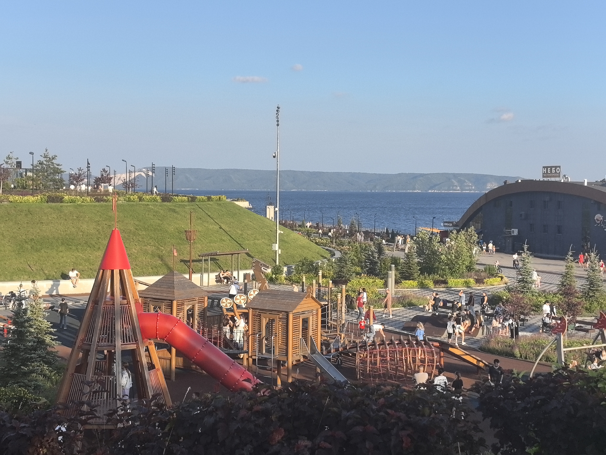 This is a wide-angle, eye-level shot of a vibrant, modern park on a sunny day. The park is situated on the bank of a large body of water, likely a river or a sea, with a distant hilly coastline visible across the water. The sky is a clear, bright blue with a few wispy clouds.
The foreground is dominated by a colorful playground with wooden structures, slides, and climbing nets. A prominent red conical tower with a spiral slide is a central feature. Several children and adults are scattered throughout the playground, engaging in various activities. Some children are playing on the equipment, while adults are supervising or socializing.
In the middle ground, a lush green grassy hill slopes down towards the water. There are pathways and benches on the hill, and more people are seen walking and resting. Beyond the hill, along the waterfront, there is a paved promenade lined with trees and streetlights, with more people enjoying the scenery.
In the background, a large, dark-colored building with a curved roof stands out. A sign on this building reads "НЕБО GASTROPOINT". Further in the distance, a bridge can be seen spanning the water.
The overall mood is lively and relaxed, suggesting a pleasant summer afternoon. The sunlight casts soft shadows, indicating it is likely late afternoon or early evening. The location appears to be a well-maintained public park designed for recreation and leisure, with clear indications of being in Tolyatti, Russia.