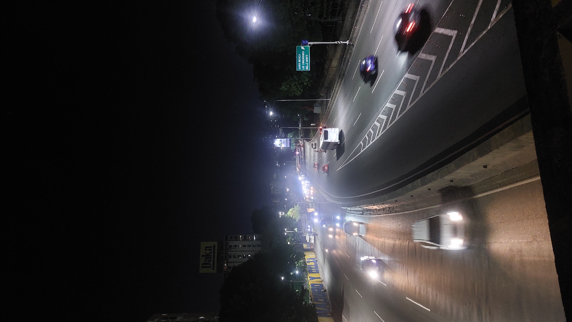 The image depicts a nighttime view of a highway in Caracas, Venezuela. The road is illuminated by streetlights and the headlights of moving vehicles, creating light trails. A building with a "Daka" advertisement is visible in the background. A sign indicates destinations such as "Santa Monica," "Los Chaguaramos," and "Bello Monte." The perspective suggests the photo was taken from an elevated position, possibly a bridge or overpass. The scene shows moderate traffic flow.