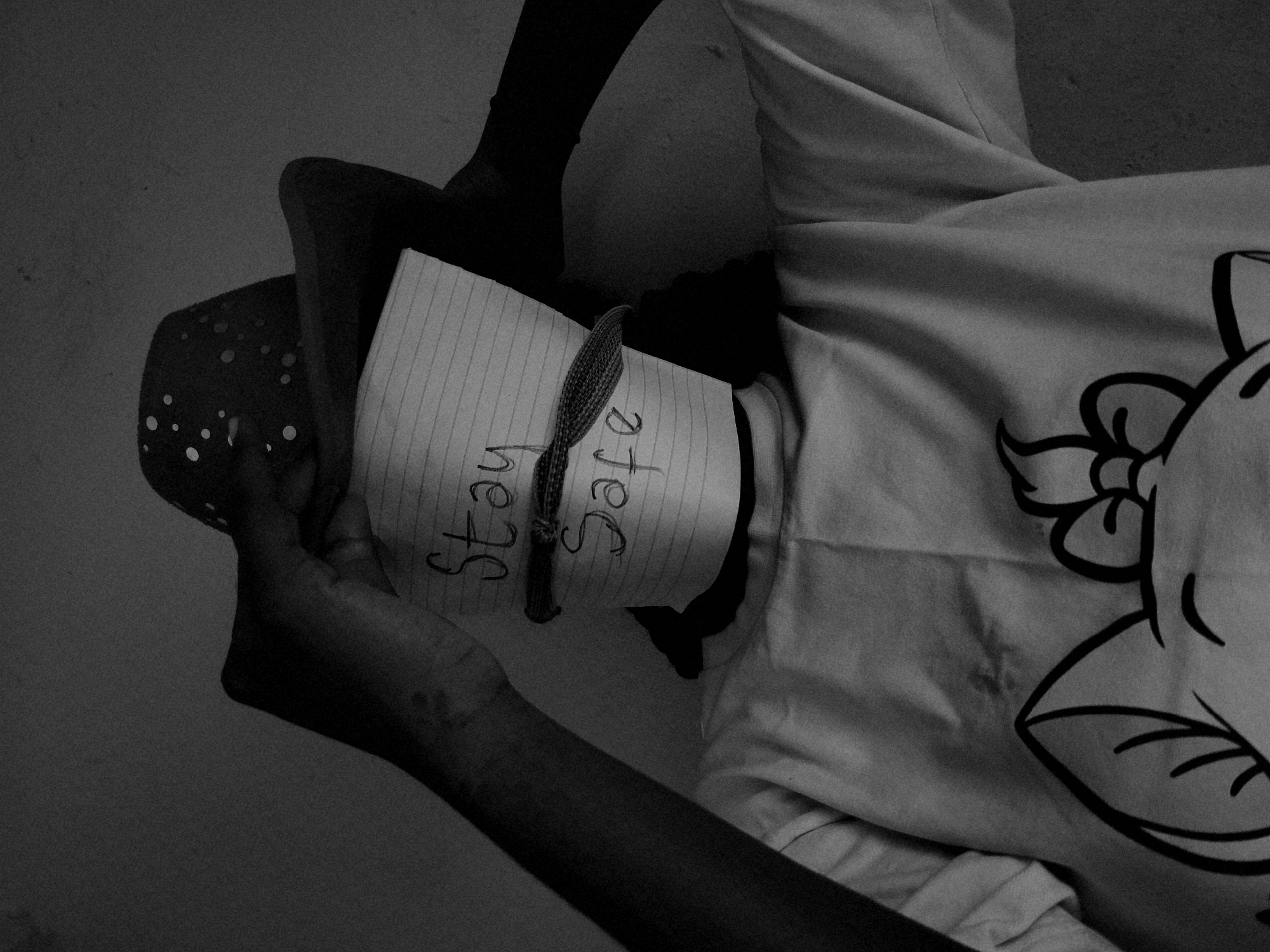 A black and white image shows a person lying down, possibly indoors, with a piece of lined paper wrapped around their head. The paper reads "Stay Safe" in handwritten text. A braided cord or string encircles the paper around the person's neck. The person is wearing a light-colored t-shirt with a cartoon flower graphic on it. A spotted or dotted fabric covers the left side of the person's face and head. The location is presumed to be Jos, Nigeria.