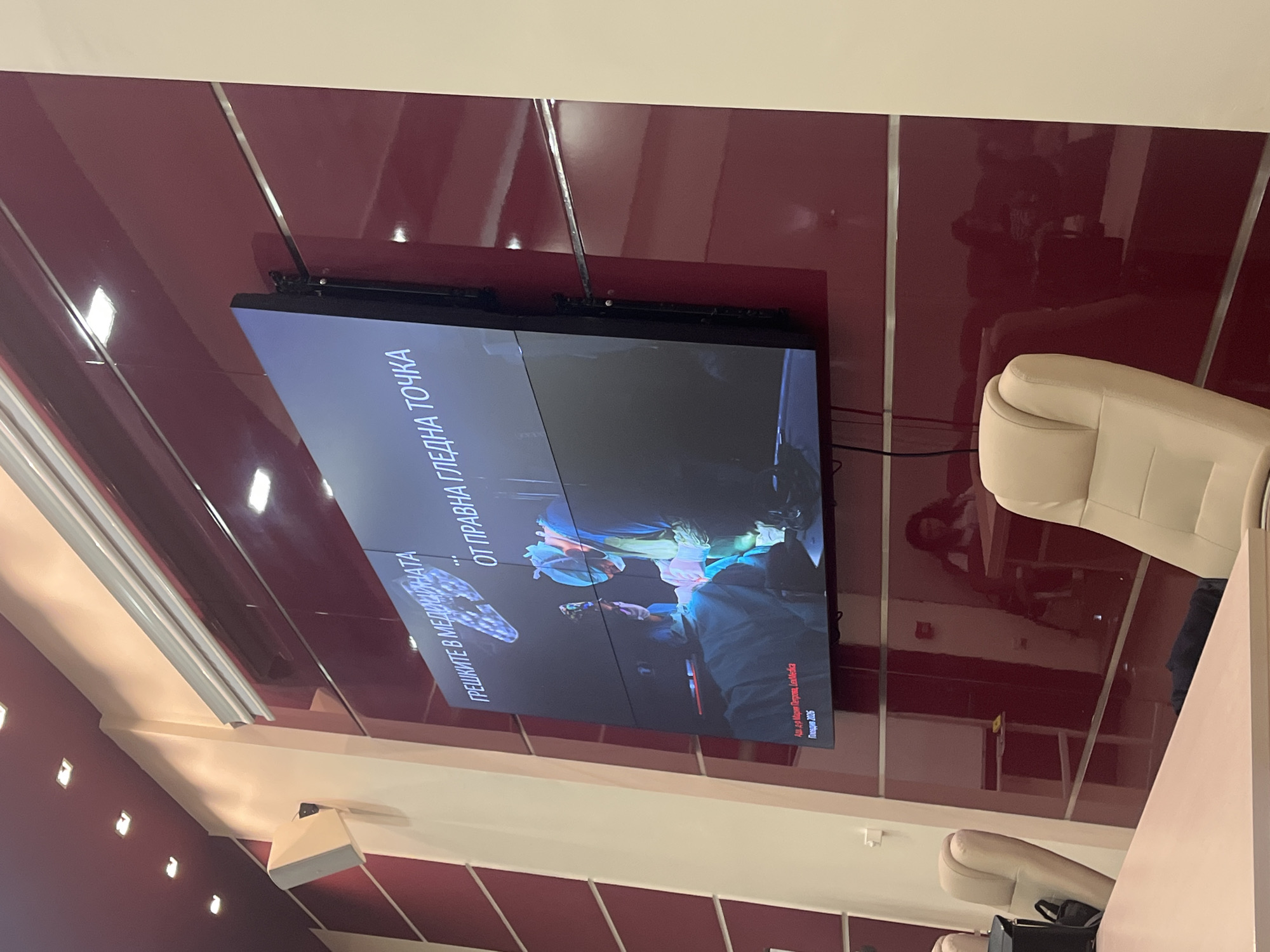 An indoor scene, possibly a conference room or lecture hall, features a large, horizontally oriented screen mounted on a glossy, dark red wall. The wall is interspersed with vertical silver or chrome strips and reflects various elements of the room, including the person taking the photo and other attendees. A cream-colored or light beige wall or ceiling section is visible above the red wall, fitted with artificial lighting. To the right of the screen, a plush, light-colored armchair is partly visible.
The screen displays a presentation in Bulgarian, with the title "ГРЕШКИТЕ В МЕДИЦИНАТА" (Mistakes in Medicine) and "ОТ ПРАВНА ГЛЕДНА ТОЧКА" (From a Legal Point of View). Below the title, an image depicts what appears to be a medical procedure, with medical personnel in scrubs and caps gathered around an operating table, reinforcing the presentation's theme. Further text on the bottom right corner of the screen reads "Асс. д-р Мария Петрова, LexMedica" (Assoc. Dr. Maria Petrova, LexMedica) and "Пловдив 2026", indicating the speaker and organization, as well as the location (Plovdiv, Bulgaria) and a possible future date for the event or presentation material. The glossy walls show multiple reflections, including what appears to be the silhouettes of people in the room. The setting suggests an educational or professional event.