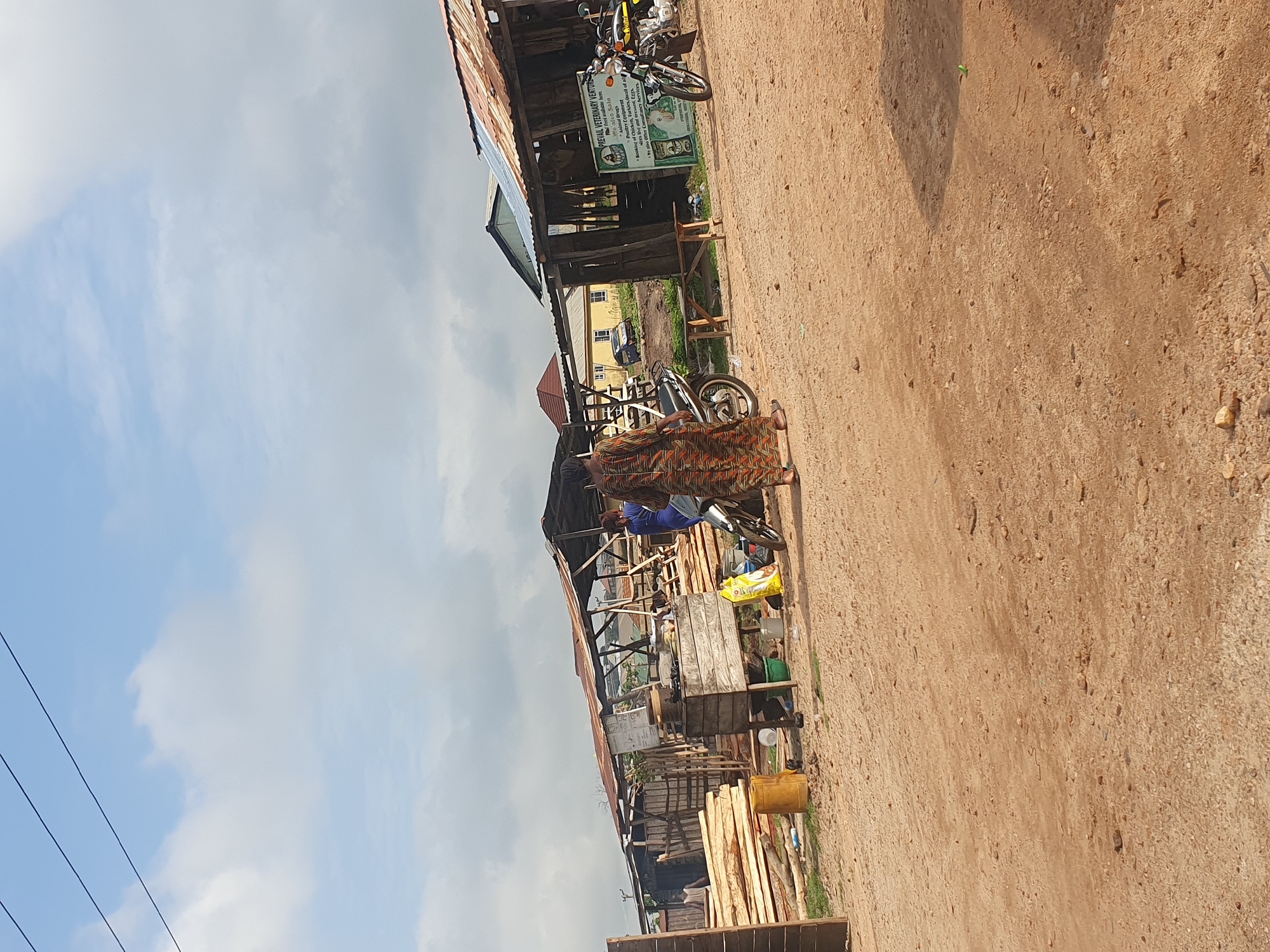The image captures a street scene in Lokoja, Nigeria. The foreground is dominated by a dirt road that stretches into the distance.
Alongside the road is a rough, wooden structure that appears to be a small business. Lumber is stacked against the structure. A sign reads "PREVAIL VETERINARY VENTURES." A motorcycle is parked nearby. A person dressed in colorful attire is walking along the side of the road near the business. The sky is partly cloudy. Overhead electrical wires are visible. A building is visible in the background behind the shop.