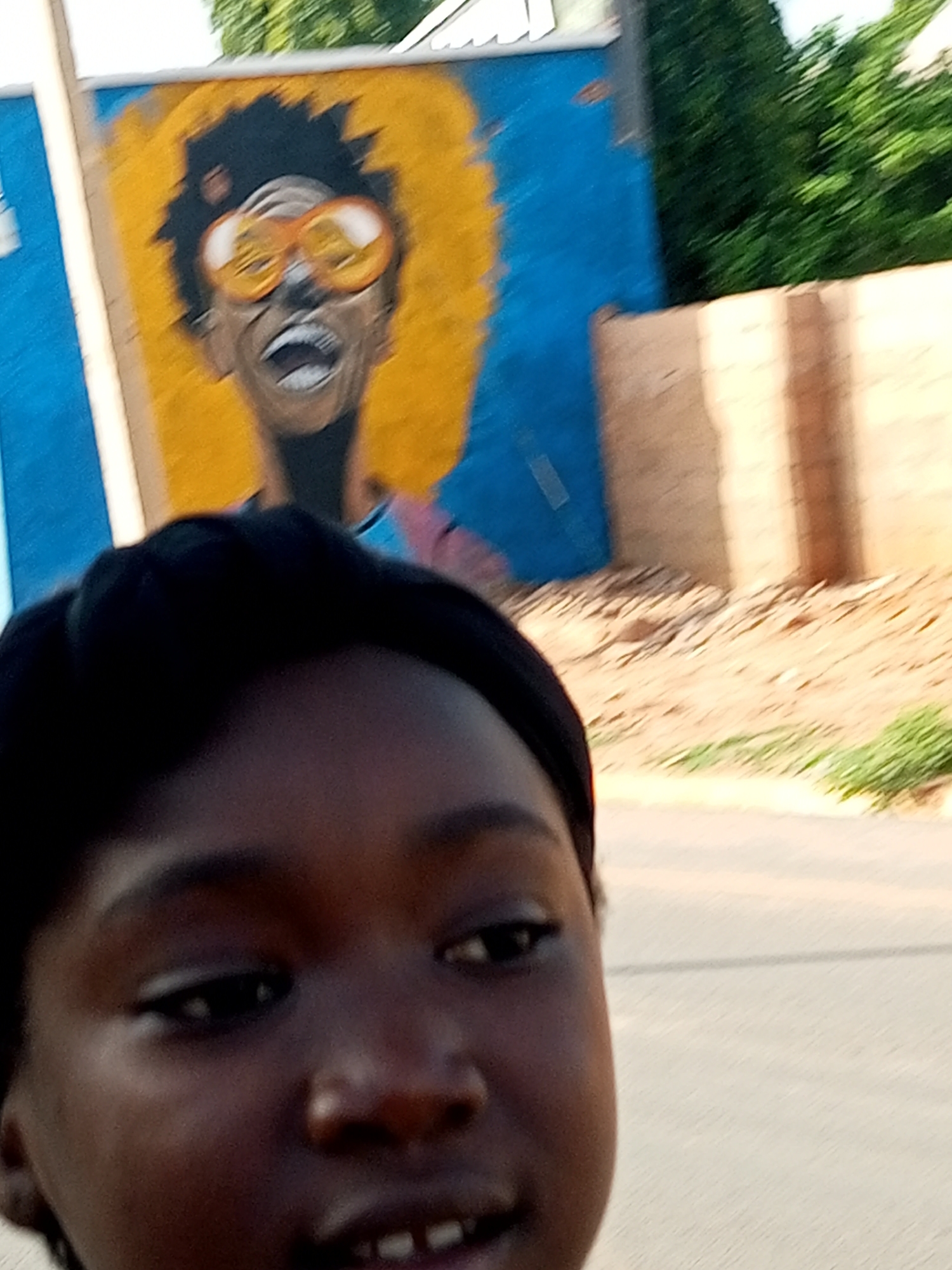 A young Black female is taking a selfie outdoors. She is positioned in the foreground, occupying the majority of the frame. Her dark hair is visible, and her facial features, including her eyes, nose, and mouth, are slightly blurred due to the close proximity to the camera. Her mouth is open, revealing teeth. In the background, a mural is depicted on a blue wall. The mural features a portrait of a person with dark, curly hair and oversized orange sunglasses, and a large yellow halo or afro. The mural is positioned on a wall, with a brick structure visible to the right. Green foliage is visible behind the mural. The setting appears to be a public space, possibly a street or an alleyway in Tempure, Nigeria. The lighting suggests it is daytime.