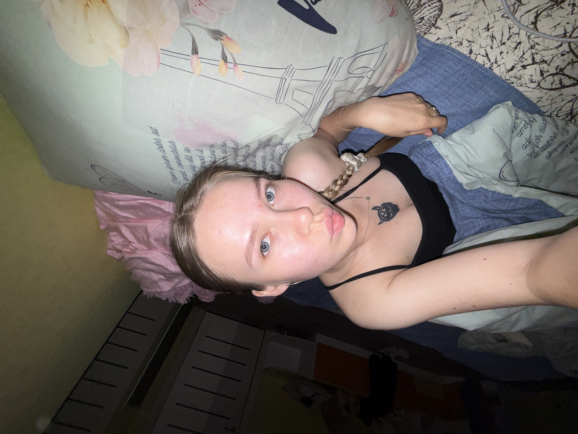 A young woman, likely in her late teens or early twenties, is captured in a selfie while lying on a bed in Zhigalki, Russia. Her head is tilted, giving the image a rotated perspective. She has blue eyes, a clear complexion with minor blemishes, and light-colored hair, part of which is styled in a braid secured with a white scrunchie. Her lips are slightly pursed in a pout as she looks directly at the camera, holding the phone with her right hand.
She is wearing a black spaghetti strap top, which reveals a black turtle-shaped pendant on a silver chain around her neck. A thin gold-colored bracelet is visible on her right wrist, and a ring adorns her right ring finger.
The setting is a bedroom. She is lying on a bed with multiple layers of bedding. A light blue-green pillow beneath her head features a pattern of soft-hued flowers and a sketch of the Eiffel Tower. This pillow also contains visible text, including phrases like "Lorem ipsum dolor sit amet, consectetur adipiscing elit," which appear to be placeholder text. Another light blue textured blanket covers part of her body. Behind her head, a fluffy pink blanket or throw is visible. The background also shows a dark, abstractly patterned bedspread, a dark furniture piece with horizontal slats, and an olive-green wall. The subdued lighting suggests an indoor shot, possibly taken in the evening or at night.