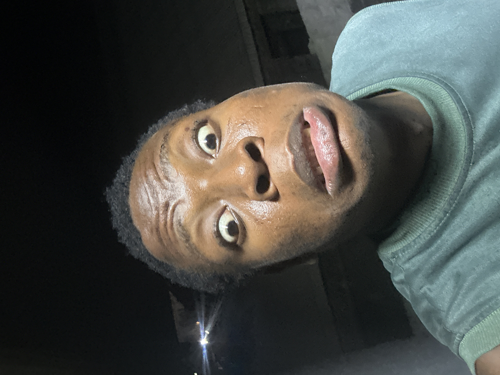 A close-up, angled, upward-facing selfie shows a dark-skinned person wearing a light green long-sleeved shirt. The person's face is the primary focus, with visible sweat. A streetlight shines in the lower left corner, suggesting an outdoor setting at night. The specific city and location are unknown.