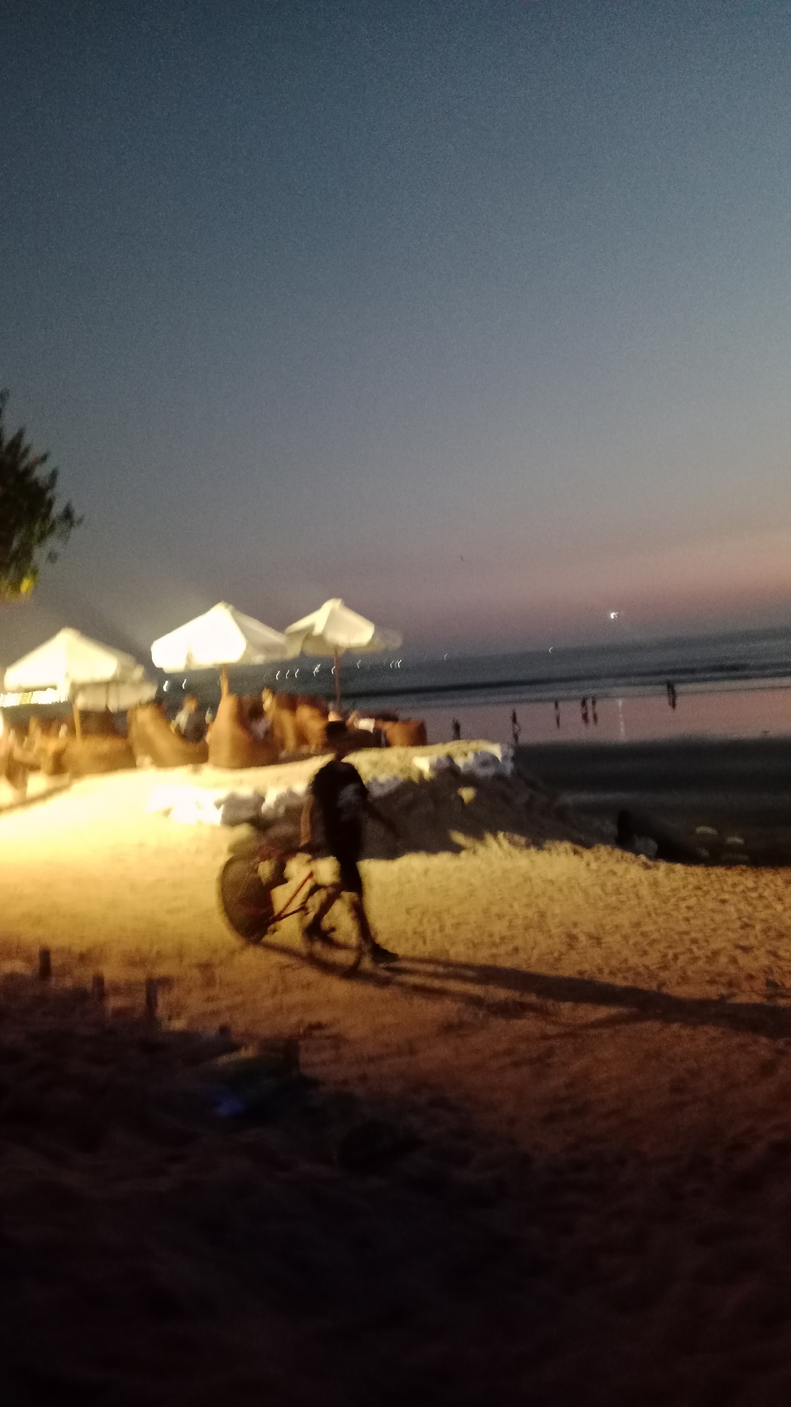 This image depicts a beach scene at dusk in Kuta, Indonesia. The sky is a gradient of dark blue to lighter shades of gray and pink near the horizon. In the foreground, the sandy beach is illuminated by artificial light, creating a warm glow. A person on a bicycle is the most prominent subject, silhouetted against the brighter sand. To the left, several white beach umbrellas are set up, with beanbag chairs and what appear to be tables beneath them, suggesting a seating area for patrons of a beachside establishment. Some figures are visible in the background, standing near the water's edge, where gentle waves are breaking on the shore. The overall impression is of a tranquil evening at a popular tourist destination.