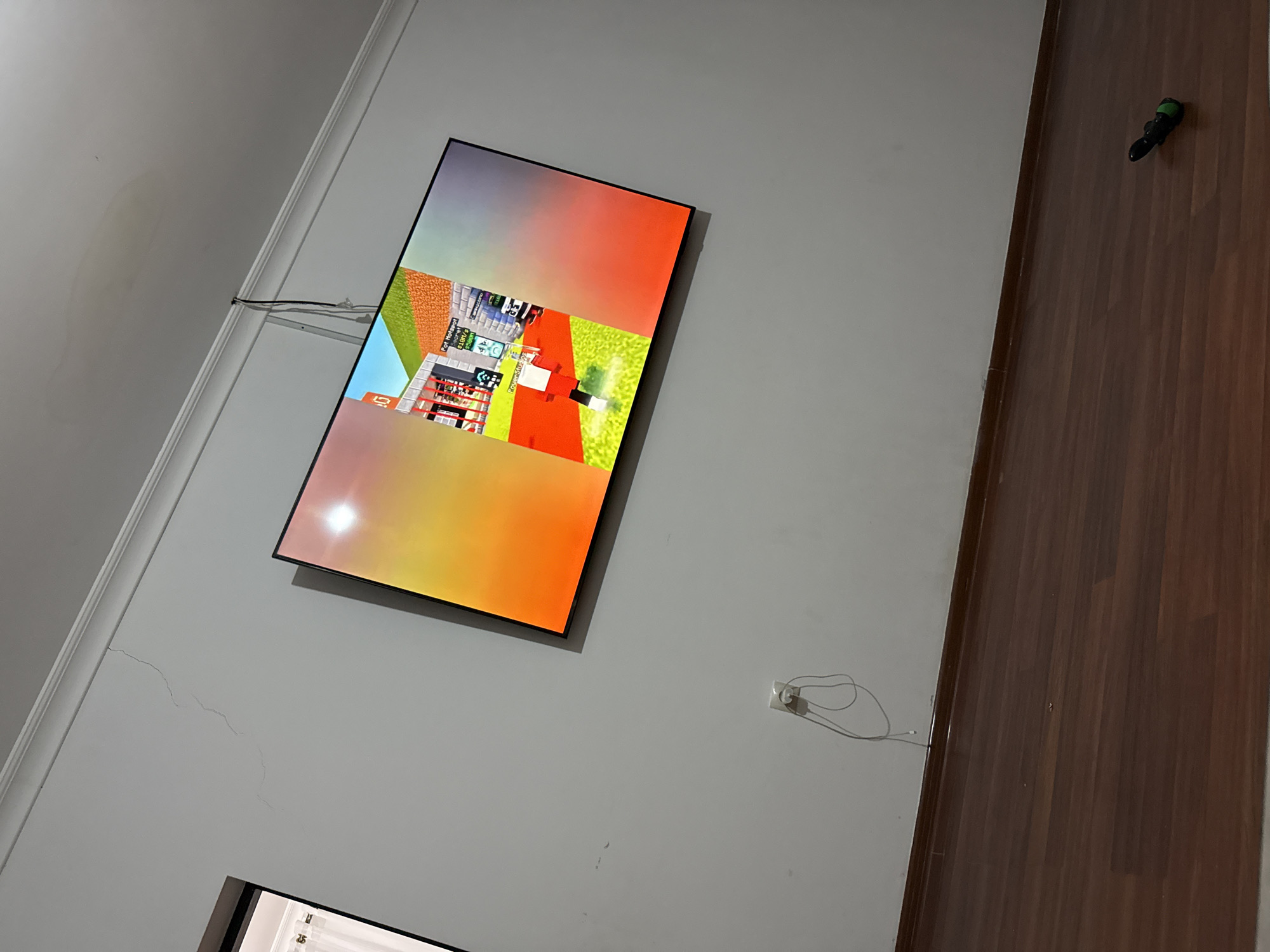 The image shows a living room with a television mounted on the wall. The television is displaying a video game, possibly Minecraft. The room has light-colored walls and a wooden floor. There is a white electrical outlet on the wall with a charger plugged in. The ceiling has a water stain. The overall scene is unremarkable and does not provide any specific information about the location being in Serebryakovka, Russia.