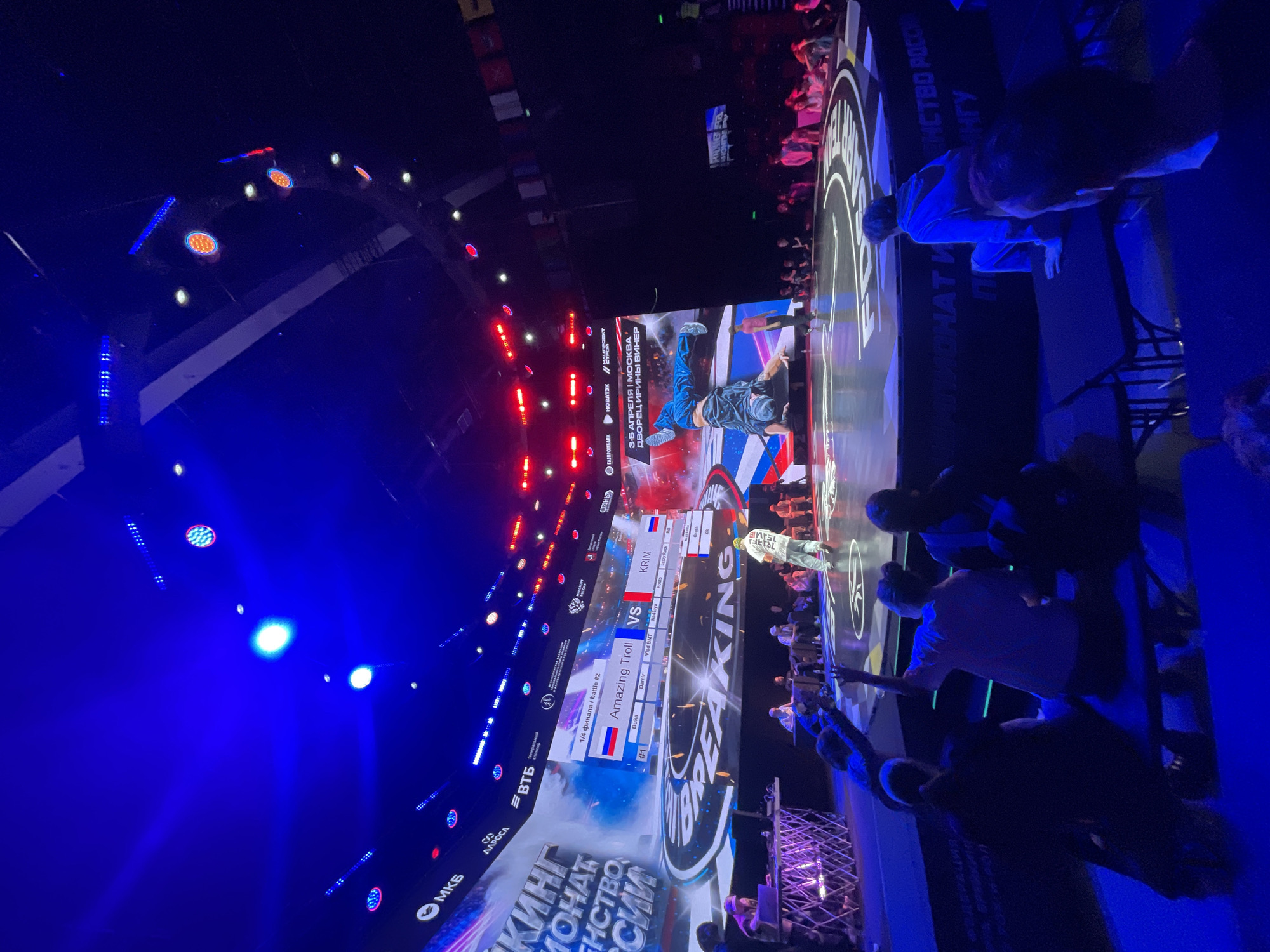 The image captures a scene at the Breaking Championship in Moscow, Russia, held between April 3-5 at the Irina Viner Palace. A screen displays a battle between Amazing Troll (Buka and Damir) and KRIM (Vlad BMT, Ka4elya, and Jazzy Rock). Spectators are present, watching the event. The scene includes the stage lighting and event branding.