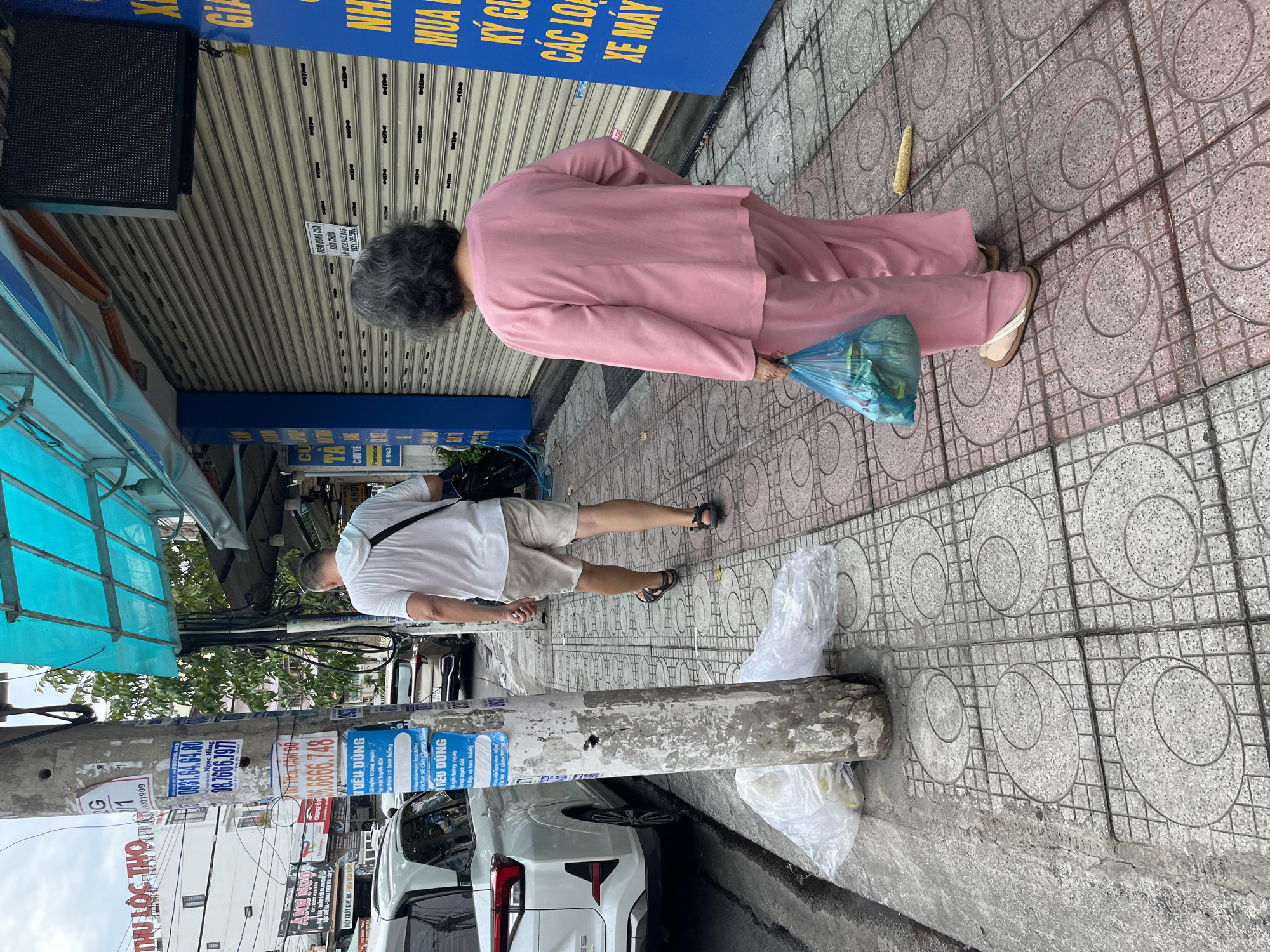 The image shows a street scene in Nha Trang, Vietnam. In the foreground, an elderly woman wearing a pink tracksuit and sandals walks away from the camera, carrying a blue plastic bag. To her left, a man in a white t-shirt and beige shorts is also walking away, with a black bag slung over his shoulder. A white car is parked on the left side of the street. A utility pole with various posters and flyers is visible in the center of the image. The sidewalk is paved with patterned tiles. In the background, there are buildings with roll-up shutters and a bright blue awning. The overall scene suggests a typical street environment in a Vietnamese city.