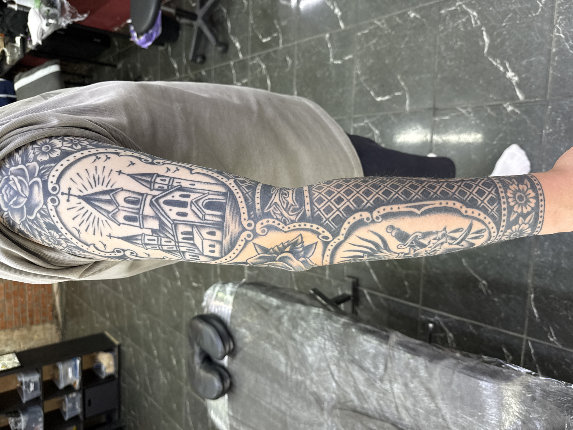 The image shows a person's tattooed arm. The tattoo is on the person's left arm and extends from the shoulder down to the wrist. The tattoo features a detailed black and grey design. The upper portion of the tattoo depicts a church with a radiating sun and birds. Below this, there are floral elements and a rose. Further down, the tattoo includes a sword with decorative elements and a geometric pattern of diamonds. The lower part of the tattoo features a floral design on the wrist. The person is wearing a light brown t-shirt and black leggings, with white socks. The background appears to be a tattoo studio, with a massage table covered in plastic wrap on the left, and shelves with containers in the upper left corner. The floor is tiled with a green and grey marbled pattern. Chairs and tables are visible in the background. The lighting suggests an indoor environment. There is no specific indication of the location being in Moscow, Russia, in the image itself.