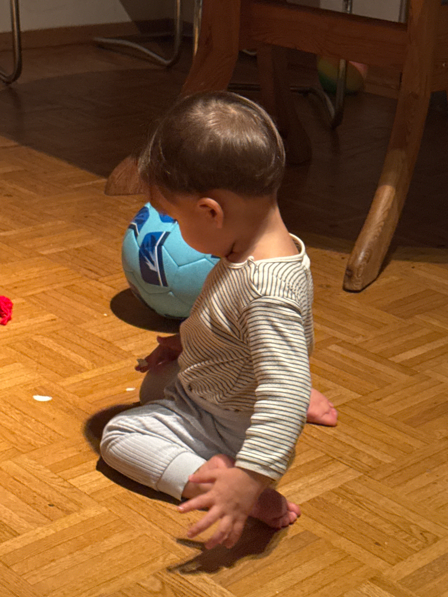A young child, seen from the side and slightly from the back, sits on a polished wooden floor with a parquet pattern. The child is wearing a long-sleeved striped shirt and light-colored pants. To the child's left is a light blue soccer ball with white and dark blue markings. A small, bright red object is visible on the floor to the far left. The scene appears to be indoors, possibly in a living area, with the leg of a wooden table or chair visible in the background. The lighting suggests it is daytime, with shadows cast on the floor. The image was captured in Überherrn, Germany.