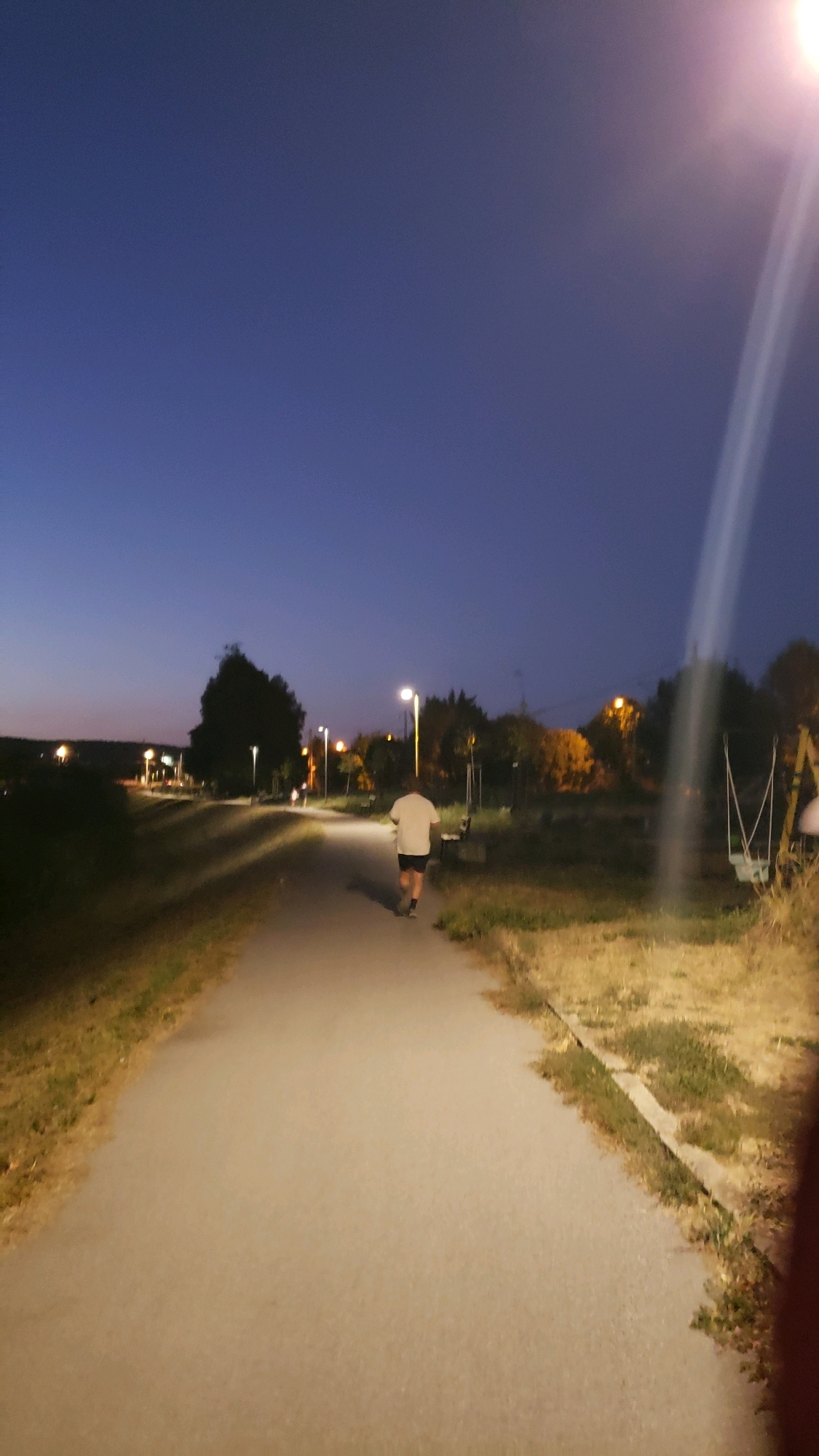 A person in a white t-shirt and dark shorts is seen from behind, running on a paved path in a park or recreational area. The sky is a deep blue, indicating either early morning or late evening. Streetlights are illuminated along the path, casting shadows and illuminating the surrounding dry grass and trees. In the background, there are more trees, some with yellow foliage, and distant lights suggest the presence of a city or town. A swing set is visible to the right of the path. The overall scene depicts a solitary person engaged in outdoor exercise at dusk or dawn in a public space. The image does not contain specific landmarks that would pinpoint the exact location within the city of Prešov, Slovakia, but the setting is consistent with urban park infrastructure.