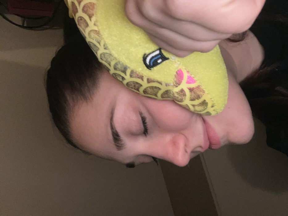 A person with closed eyes is upside down. They are holding a yellow, plush toy shaped like a snake against their face. The toy has a pattern of brown scales and a small, embroidered eye. The person's dark hair is visible behind the toy. The background is a light brown wall with a faint door or frame visible. The lighting is soft and muted.