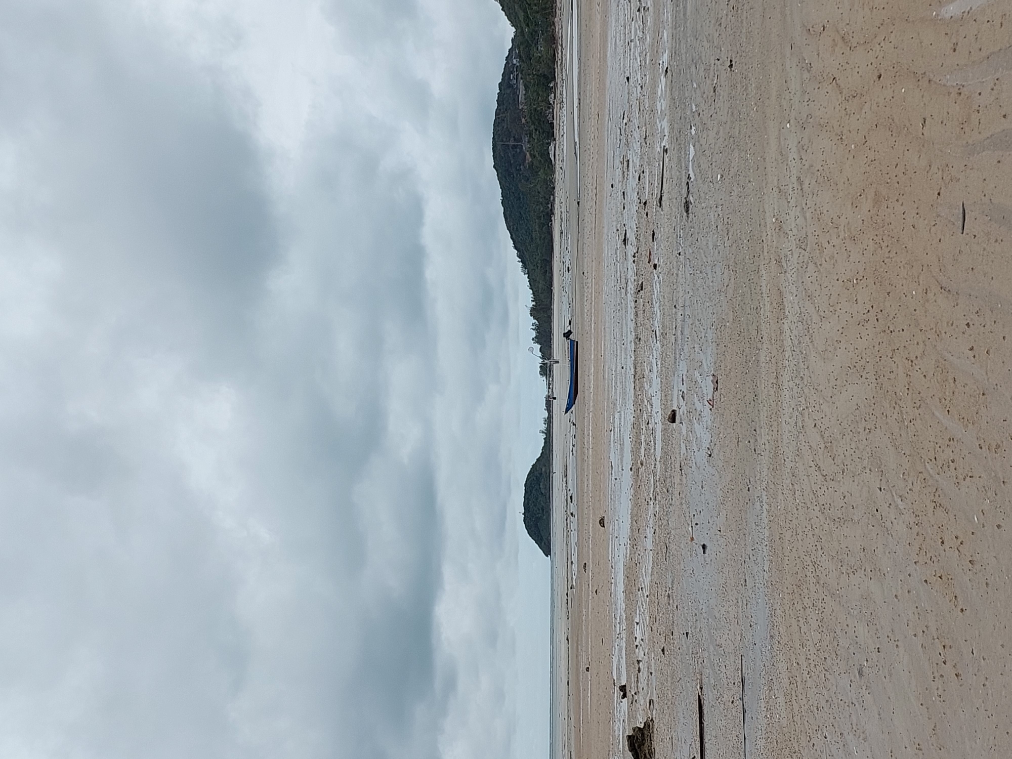 An overcast day frames a low-tide scene on a sandy beach in Ko Samui District, Thailand. The foreground is dominated by wet, sandy terrain, dotted with small shells and debris, with shallow pools of water reflecting the gray sky. In the middle ground, a lone blue and white boat rests on the sand, its engine visible. Further in the distance, two islands emerge from the sea. The closer island is small and densely forested, while the larger island in the background is a more substantial, tree-covered hill with structures and vegetation visible on its slopes. The horizon line is low, emphasizing the vastness of the cloudy sky.