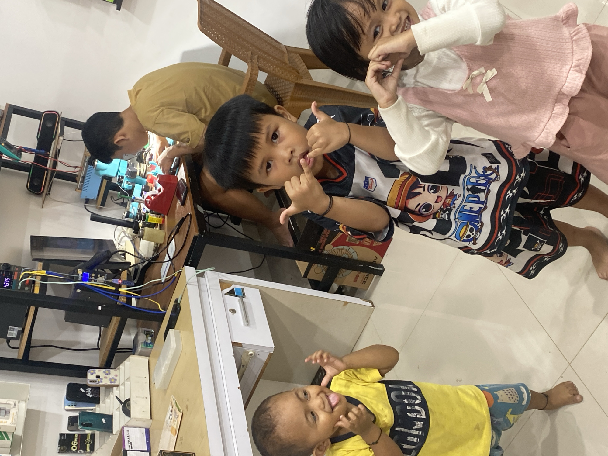 In Ta', Indonesia, three young children are posing for a photo in what appears to be a repair shop. The child on the left, wearing a yellow shirt and patterned shorts, sticks out their tongue and makes a face. In the center, a boy in a black and white shirt with a character print makes a "rock on" gesture with both hands. To the right, a girl in a pink dress over a white shirt forms a heart shape with her hands over her eyes. In the background, a young man is seated at a desk, working on electronic equipment. Shelves with various items, including what look like phone cases and electronic components, are visible. The environment is cluttered but organized, suggesting a functional workspace.