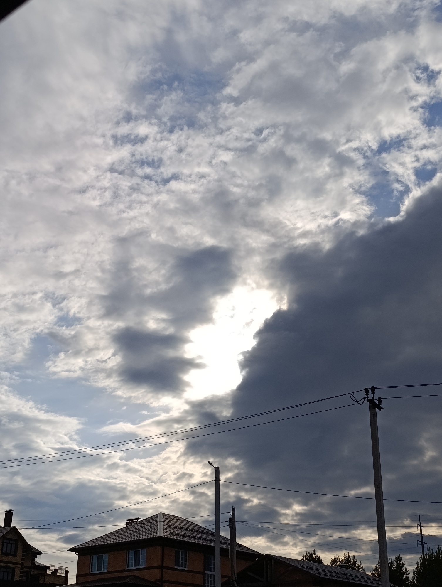 The image presents a dramatic sky filled with a dense array of clouds above a residential landscape. The upper portion of the frame is dominated by heavy, dark grey clouds, dramatically contrasting with a brilliant, luminous patch of sunlight breaking through a central opening in the cloud cover. Lighter, more diffused clouds are also visible, suggesting a dynamic and overcast weather condition.
In the lower third of the image, the upper sections and roofs of two residential houses are visible. The house on the right features a distinctive brown tiled roof with a patterned surface and appears to be constructed of brick, with glimpses of white-framed windows. To its left, another house is partially visible with a darker roof. Several utility poles, made of concrete, stand prominently, supporting numerous electrical wires that traverse the sky horizontally, adding an element of urban infrastructure to the natural backdrop. Dark green foliage, likely the tops of trees, can be seen beyond the rightmost house.
The time of day appears to be daytime, possibly late afternoon or early evening, given the strong directional light from the sun piercing the clouds. The scene conveys a sense of changing weather, with significant cloud cover but also moments of bright illumination. No people or visible text are present. The setting depicts a typical residential area, consistent with a location like Chaikovsky, Russia, blending natural sky elements with human-built structures and utilities.