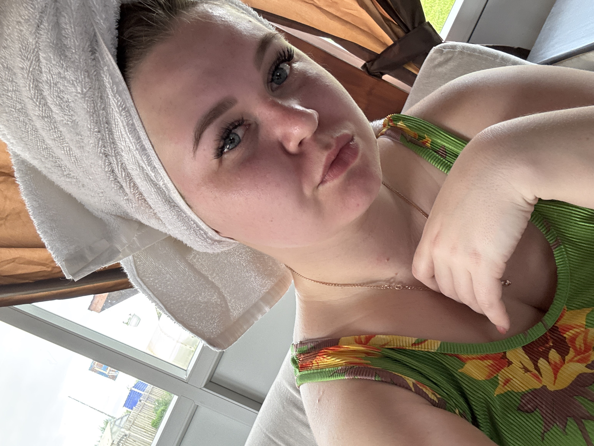 A fair-skinned woman, estimated to be in her 20s to 30s, is depicted in a close-up, selfie-style photograph taken indoors in the city of Фоки, Russia. She wears a white towel wrapped around her head and a green, ribbed tank top featuring a pattern of yellow, orange, and brown flowers. A thin gold chain with a small pendant is visible around her neck. Her left hand, with light pink nail polish, is positioned with fingers resting near her upper chest. Her face exhibits a slightly ruddy complexion, blue eyes with prominent eyelashes, and well-defined eyebrows. She looks directly towards the camera with a neutral expression. She appears to be seated or reclining on a light-colored, upholstered furniture piece. To her left, a multi-pane window with white frames provides a view of an outdoor scene under an overcast sky. Visible through the window are a wooden fence, several distant structures including a blue building, a partial view of a greenhouse, and green vegetation, indicative of a rural or semi-rural setting. A brown curtain is visible on the right side of the image.