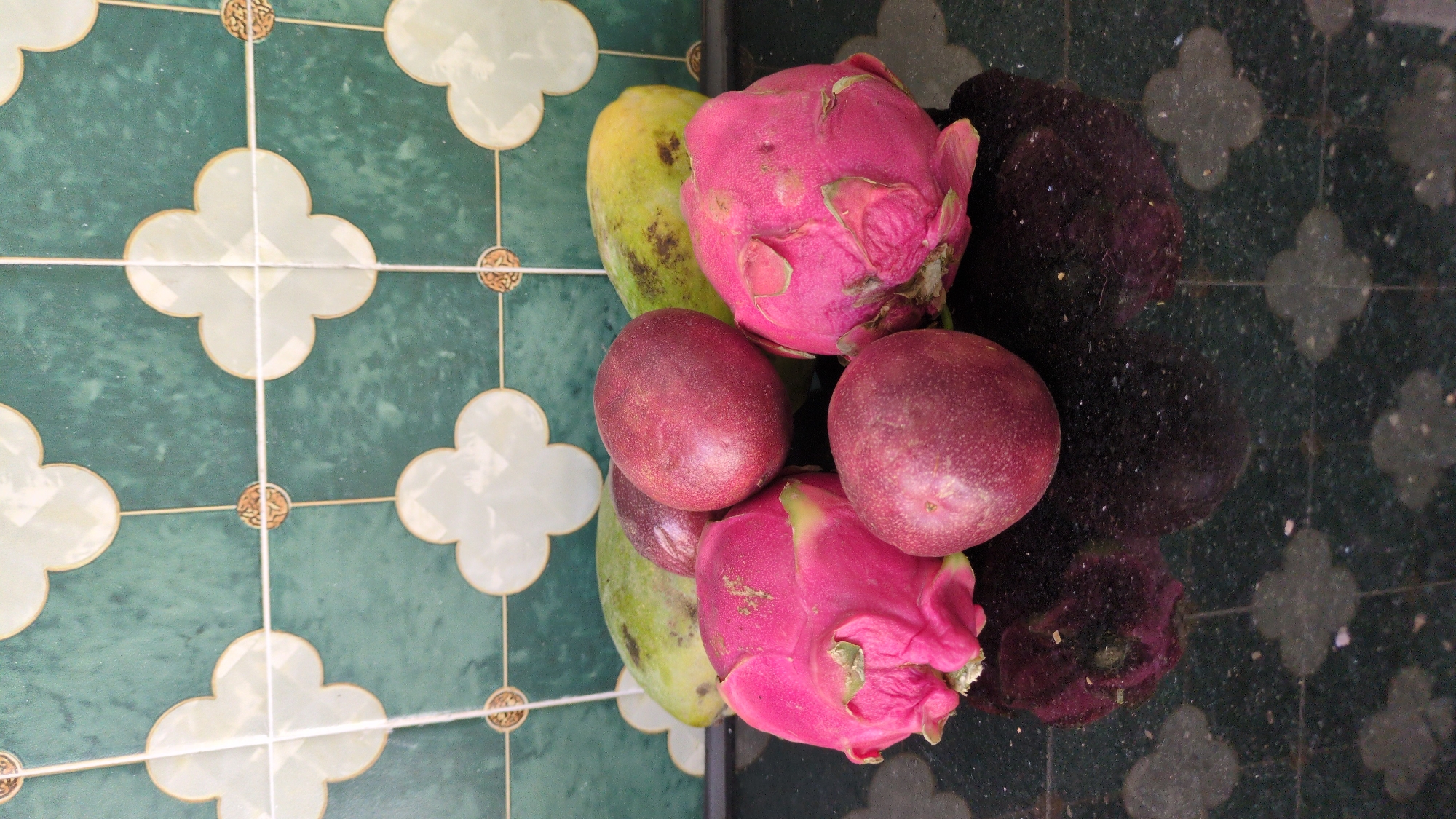 The image shows an arrangement of fruits on a tiled surface. There are two dragon fruits, a mango, and two passion fruits. The tiles are decorated with a clover pattern and the surface is glossy, reflecting the fruits. The location is identified as Da Lat, Vietnam.