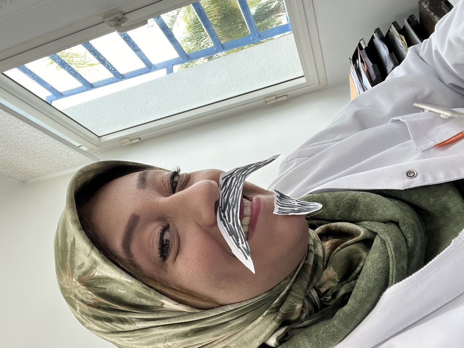 A medium close-up photograph depicts a woman in an indoor setting located in Meknes, Morocco. The subject is a woman with medium skin tone, partially visible light brown hair, and dark eye makeup, wearing a green patterned headscarf and a white lab coat with snap buttons. A red pen and a white object are clipped into the lab coat's breast pocket. Two pieces of cut paper, drawn to resemble a black-and-white striped mustache and a matching goatee, are affixed to her upper lip and chin, respectively. She is smiling, revealing upper and lower teeth, and looking directly at the camera with a playful expression. To the right of the subject, a white-framed window is visible, through which a bright sky, blue metal railings, and green palm foliage are discernible. Below the window, a stack of folders or documents is present on a surface. The interior wall is white, with a subtle white patterned texture visible above the window.