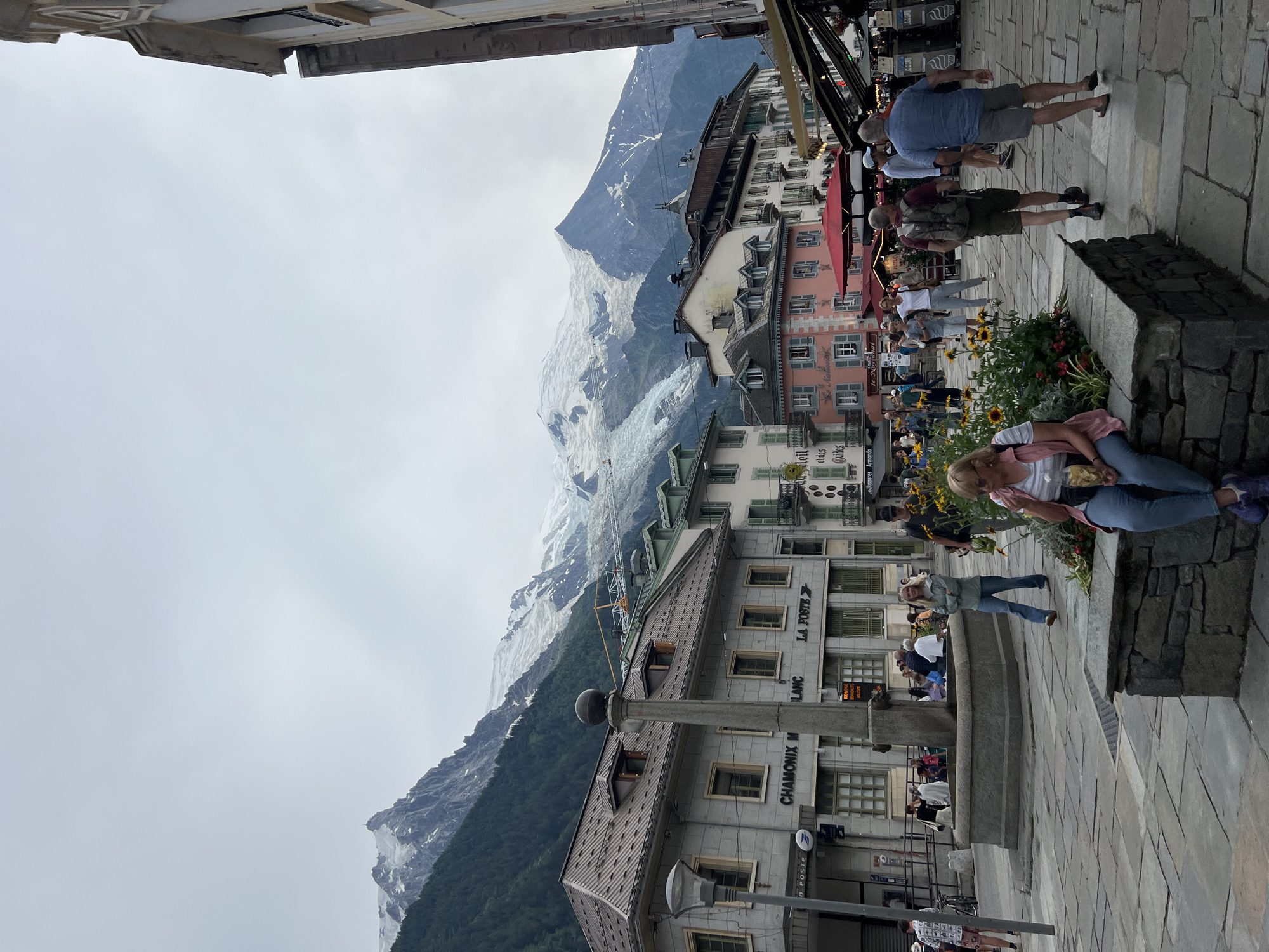 The image depicts a street scene in Chamonix-Mont-Blanc, France, with a prominent backdrop of snow-capped mountains. In the foreground, a cobblestone plaza is occupied by several people. To the left, a stone monument or fountain stands. Buildings with traditional architecture line the street, housing shops and businesses with visible signage, including "LA POSTE" and "CHAMONIX M BLANC". A group of tourists is scattered across the plaza, some walking, others seated on a stone planter box, and one person is standing near the fountain. The sky is overcast, and the overall atmosphere suggests a temperate day. The majestic mountains, likely Mont Blanc and its surrounding peaks, dominate the upper portion of the image, indicating the location's iconic natural setting.
