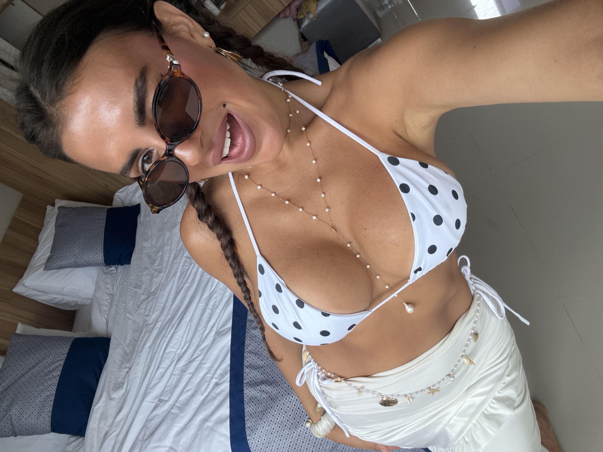 A woman poses indoors. She wears a white bikini top with black polka dots, a white skirt, sunglasses, and jewelry. The location is likely indoors, potentially a hotel room or residence. The image has a personal and candid feel. Based on the prompt it is supposedly in Kuta Selatan, Indonesia. The scene includes a bed with pillows and a blanket, part of a window, and a bare floor.
