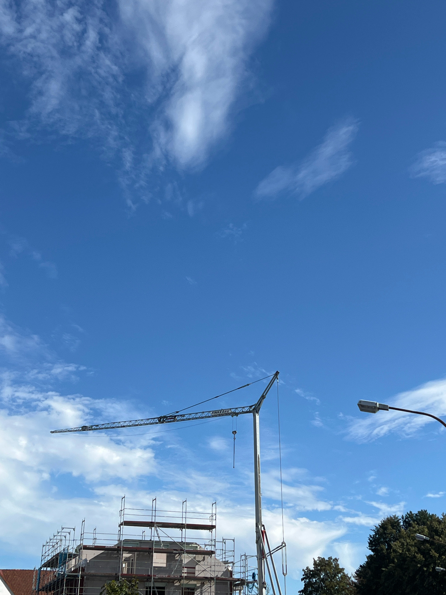 A clear, bright blue sky with scattered white clouds dominates the upper portion of the image. A construction crane with a long, horizontal boom is positioned in the middle ground, extending from right to left. The crane is anchored to a central mast, which is obscured by scaffolding. Beneath the crane, a building under construction is visible, covered in scaffolding. The building appears to be several stories high, with exposed concrete and brickwork. A street lamp with a modern design is mounted on a pole to the right of the crane, its light fixture facing downwards. In the lower right corner, the dark green foliage of mature trees can be seen. The overall scene suggests an active construction site in an urban or suburban setting.